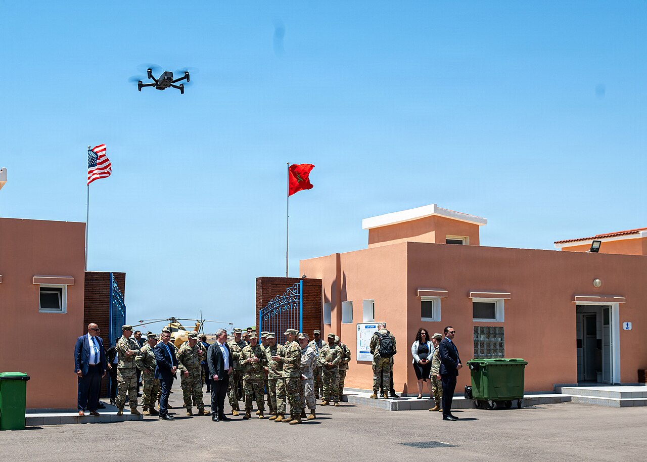 US and Moroccan officers and officials stand outside a building flying the US and Moroccan flags while a small drone hovers overhead