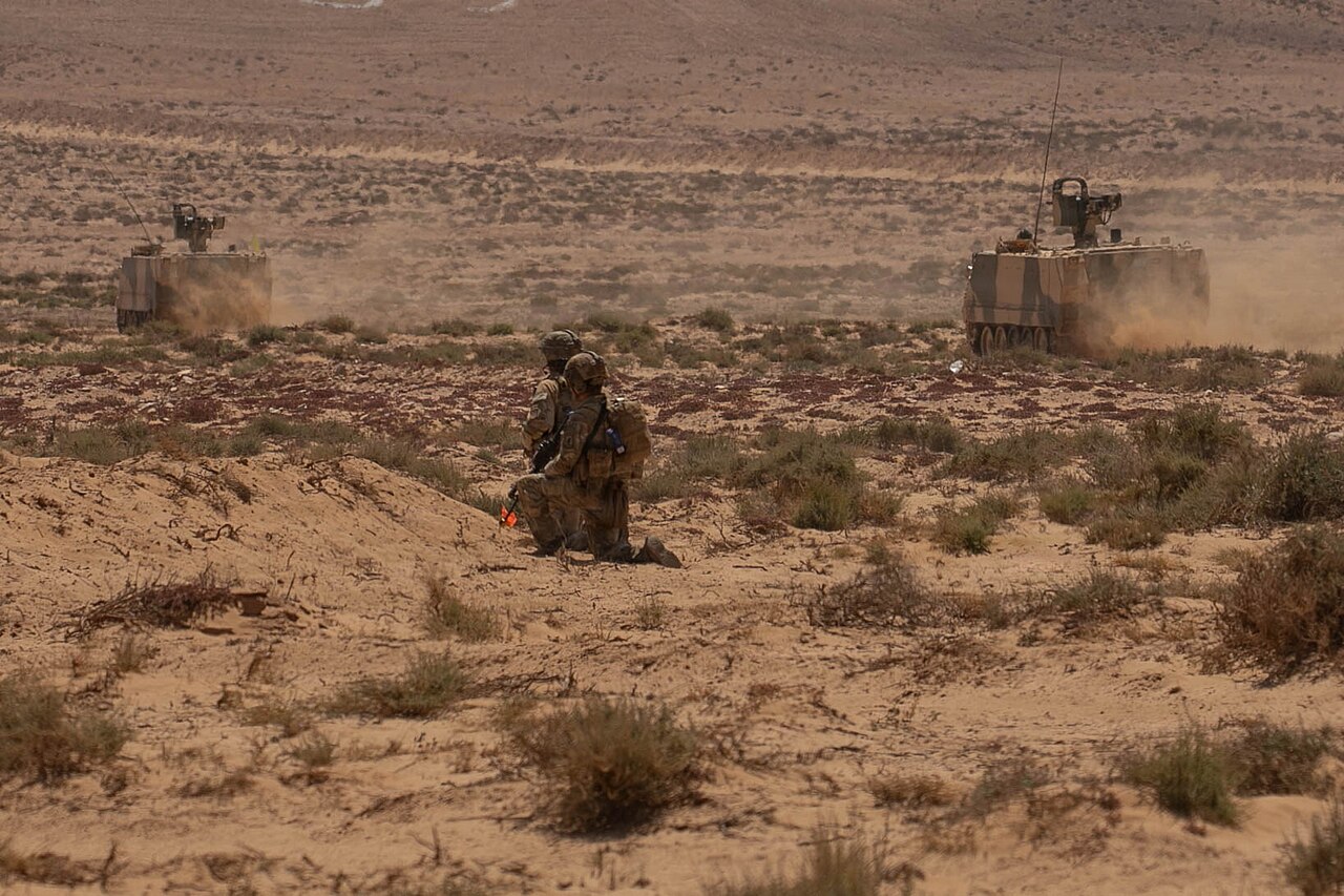 US soldiers kneel on a sandy berm as two desert-camouflaged Moroccan armoured personnel carriers advance across scrubland