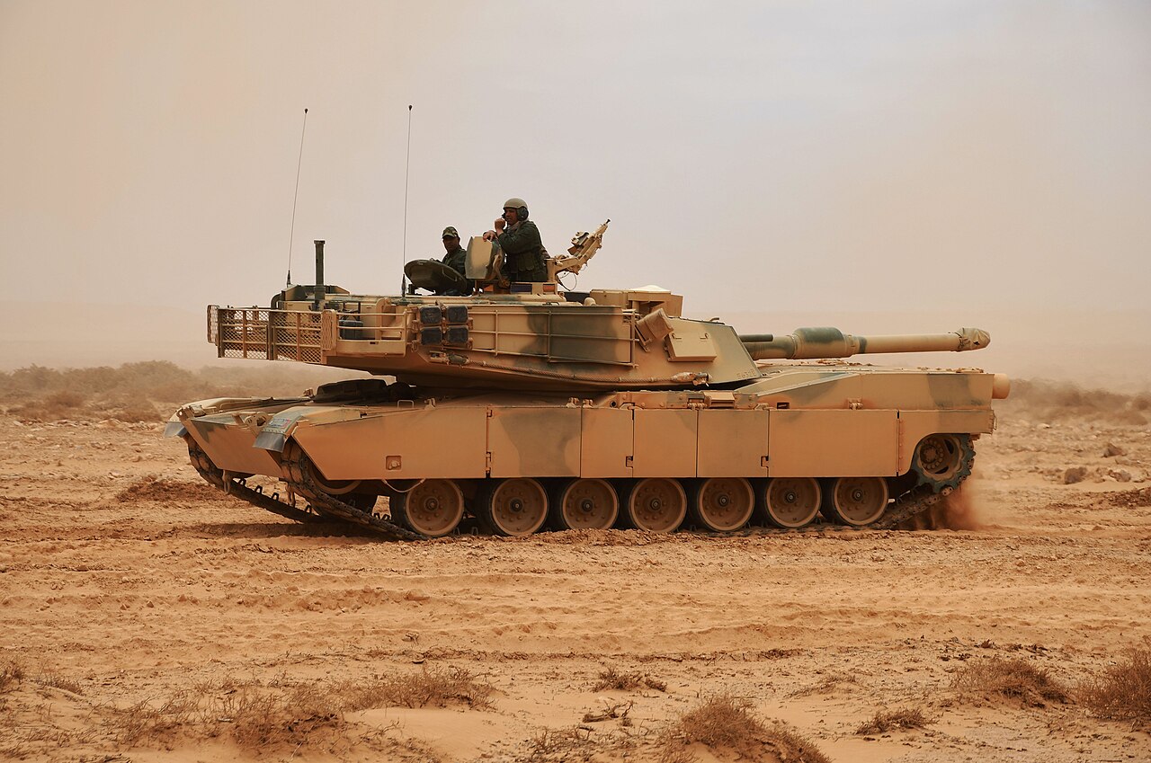 A sand-coloured Abrams tank with two crew members in the turret drives across dusty desert ground under a hazy sky