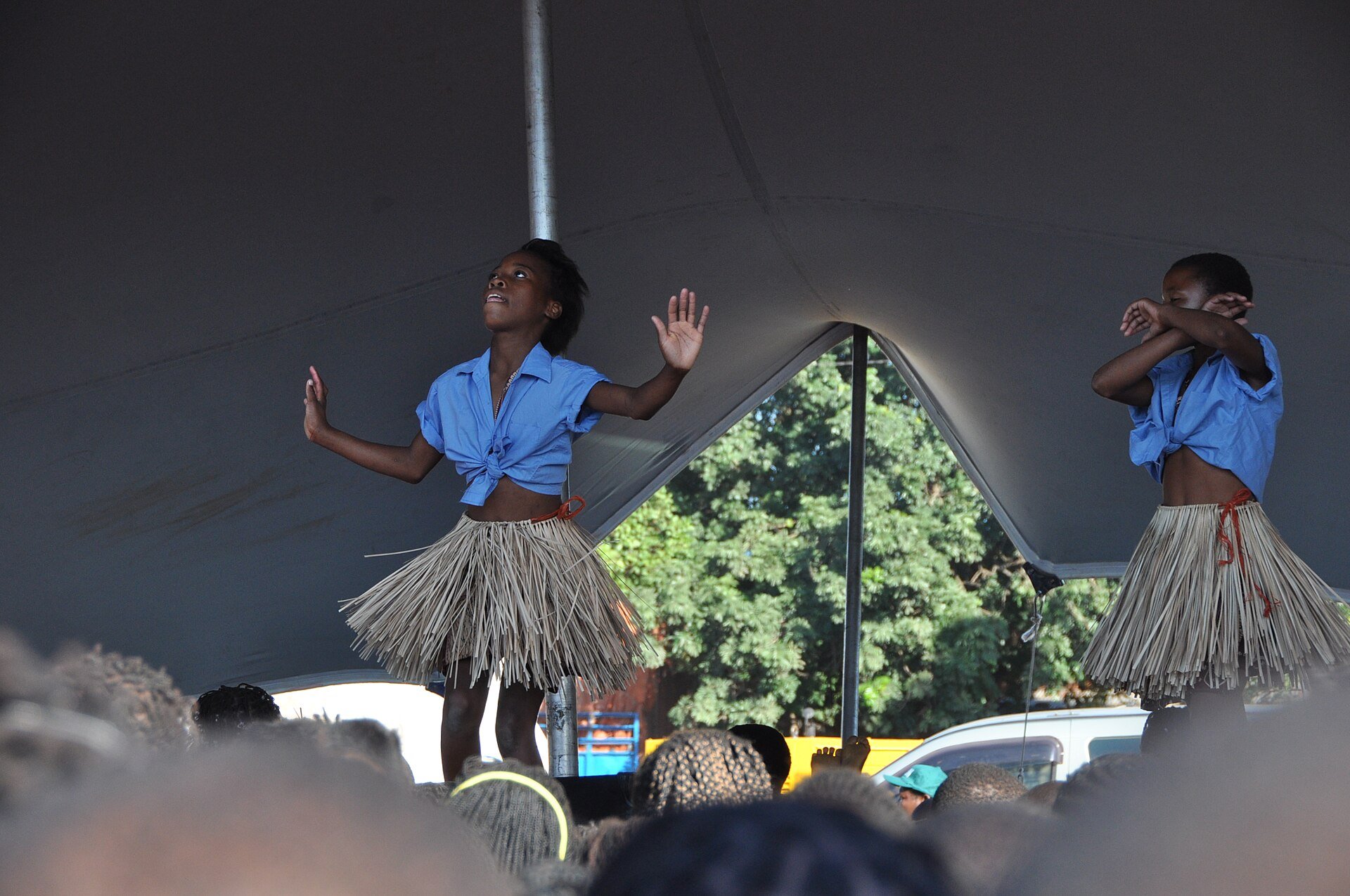 Two girls in blue shirts and raffia skirts dance on a stage under a tent in front of a crowd in Mozambique