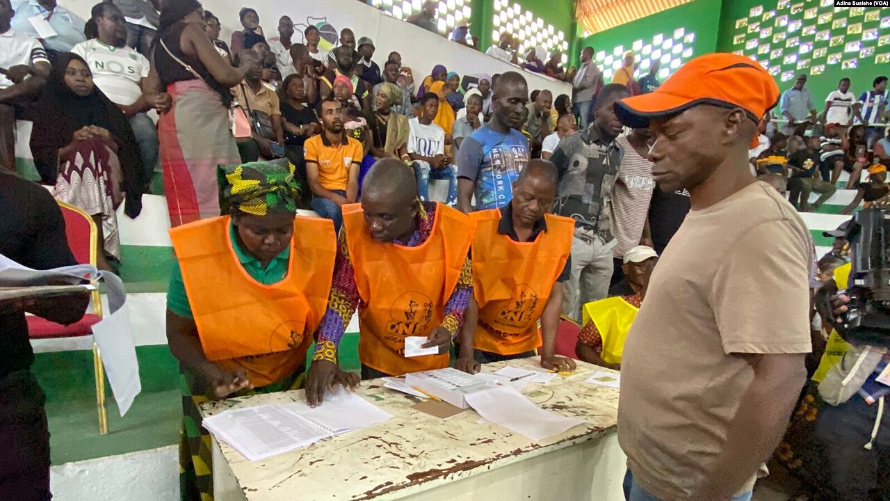 Election officials in orange vests count and record ballots at a table in a sports hall in Nampula while a man in an orange cap watches.