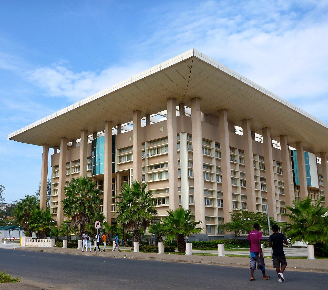 A large pale government building with a wide flat roof and tall columns, surrounded by palm trees, with pedestrians in the street in front.