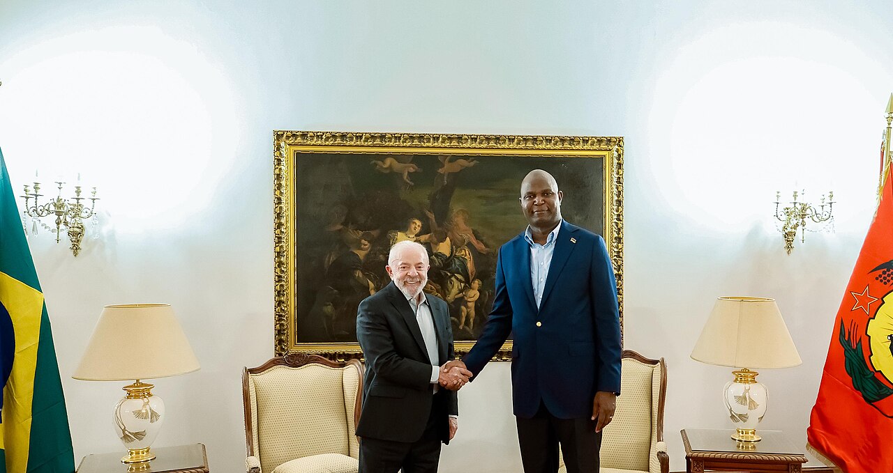 Two men in suits shake hands in a formal room between the flags of Brazil and Mozambique.