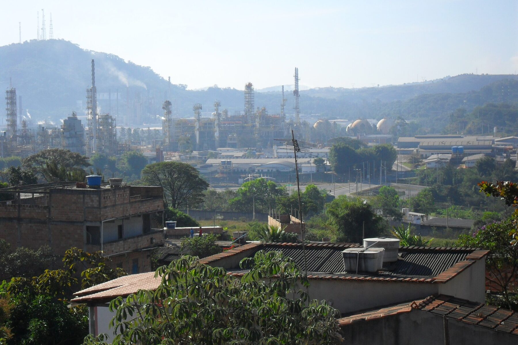 Refinery towers, pipes and storage tanks on a hazy hillside beyond houses and rooftops in Ibirité, Minas Gerais