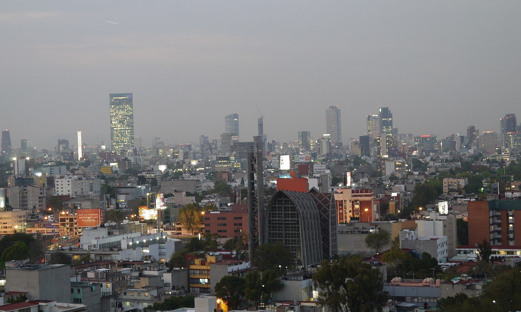 Mexico City skyline at dusk with office towers on Paseo de la Reforma rising above low-rise districts and a gray sky