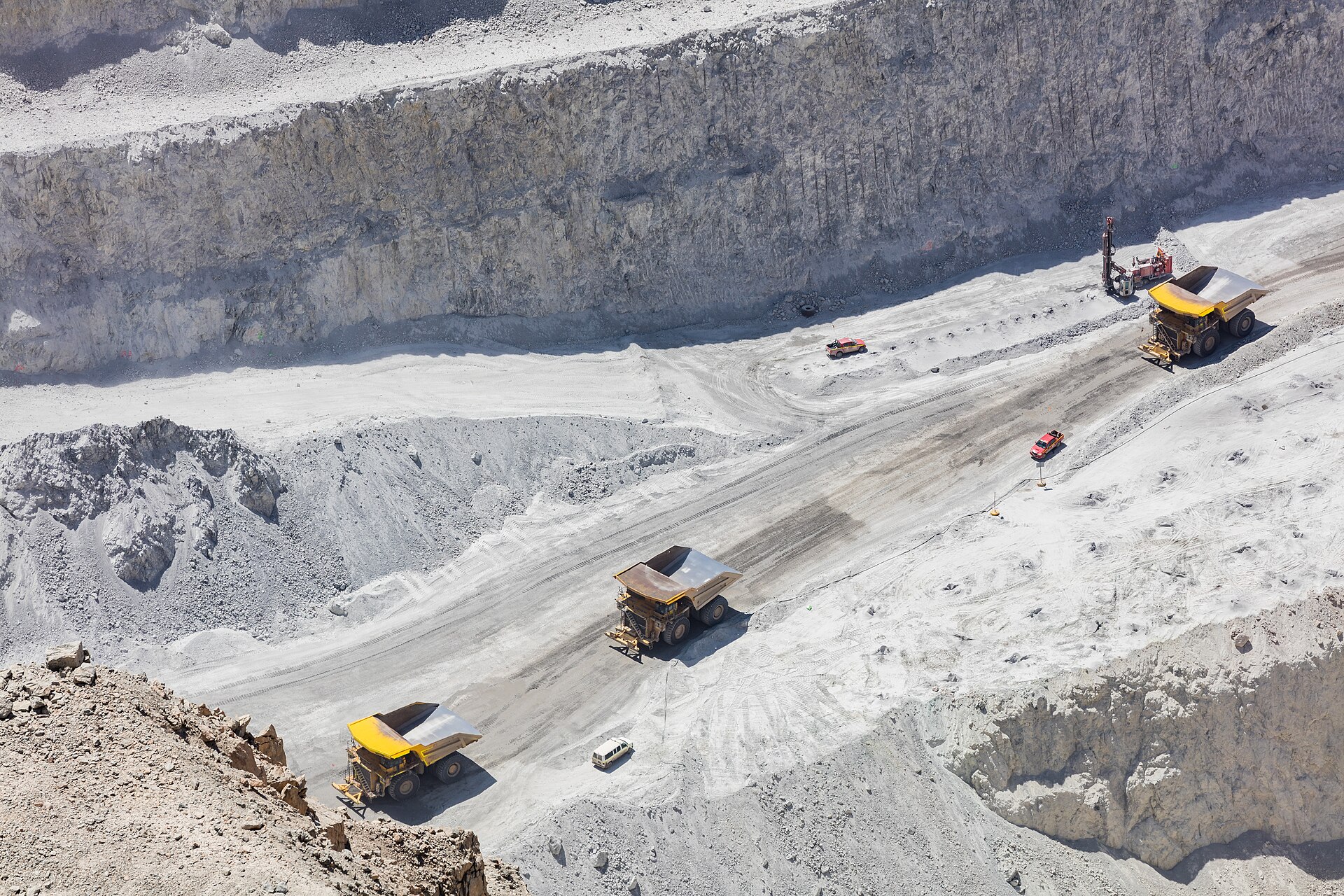 Yellow haul trucks drive along a pale grey benched road inside the Chuquicamata open pit, with a drill rig and small vehicles nearby