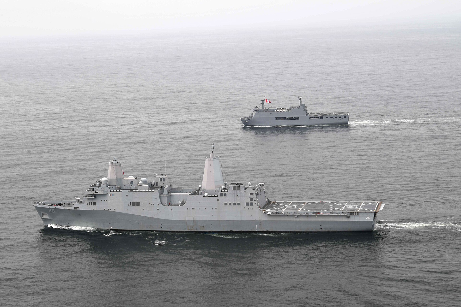 Two grey navy ships at sea, a large US amphibious ship in the foreground and a smaller Peruvian ship behind under a hazy sky