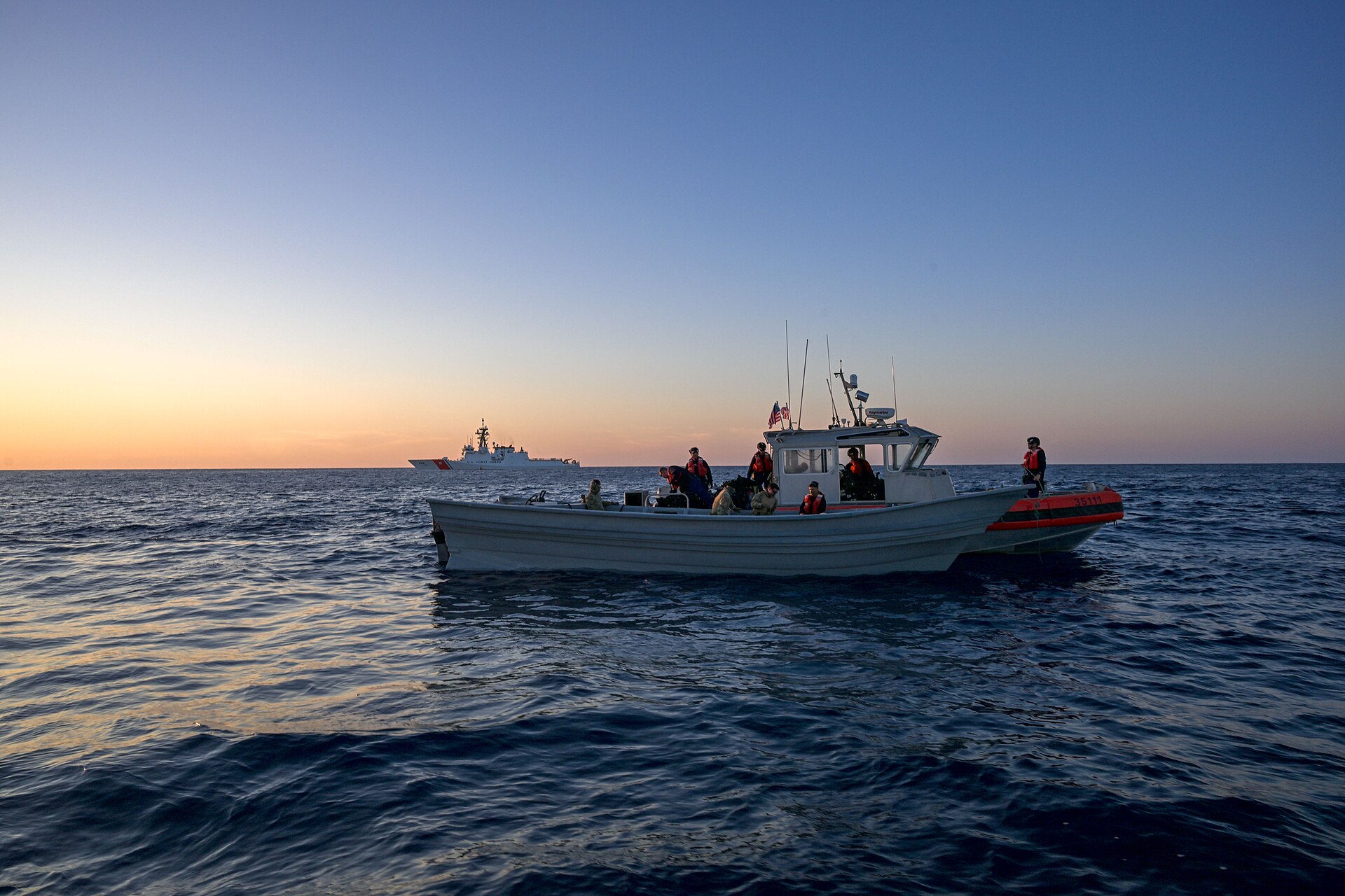 A small white boat with an orange-and-black inflatable collar carrying crew in life vests under a US flag at dusk, with a Coast Guard cutter on the horizon