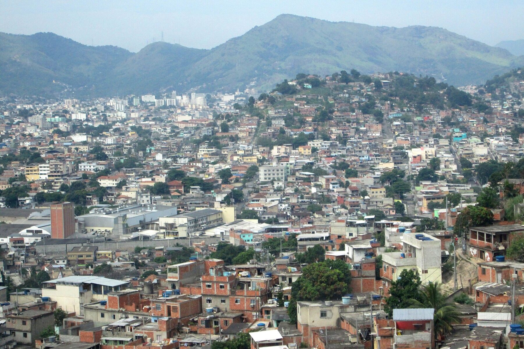 Hillside view of densely packed red-brick and concrete houses of the Complexo do Alemão favela in north Rio de Janeiro, with green hills and a hazy sky behind