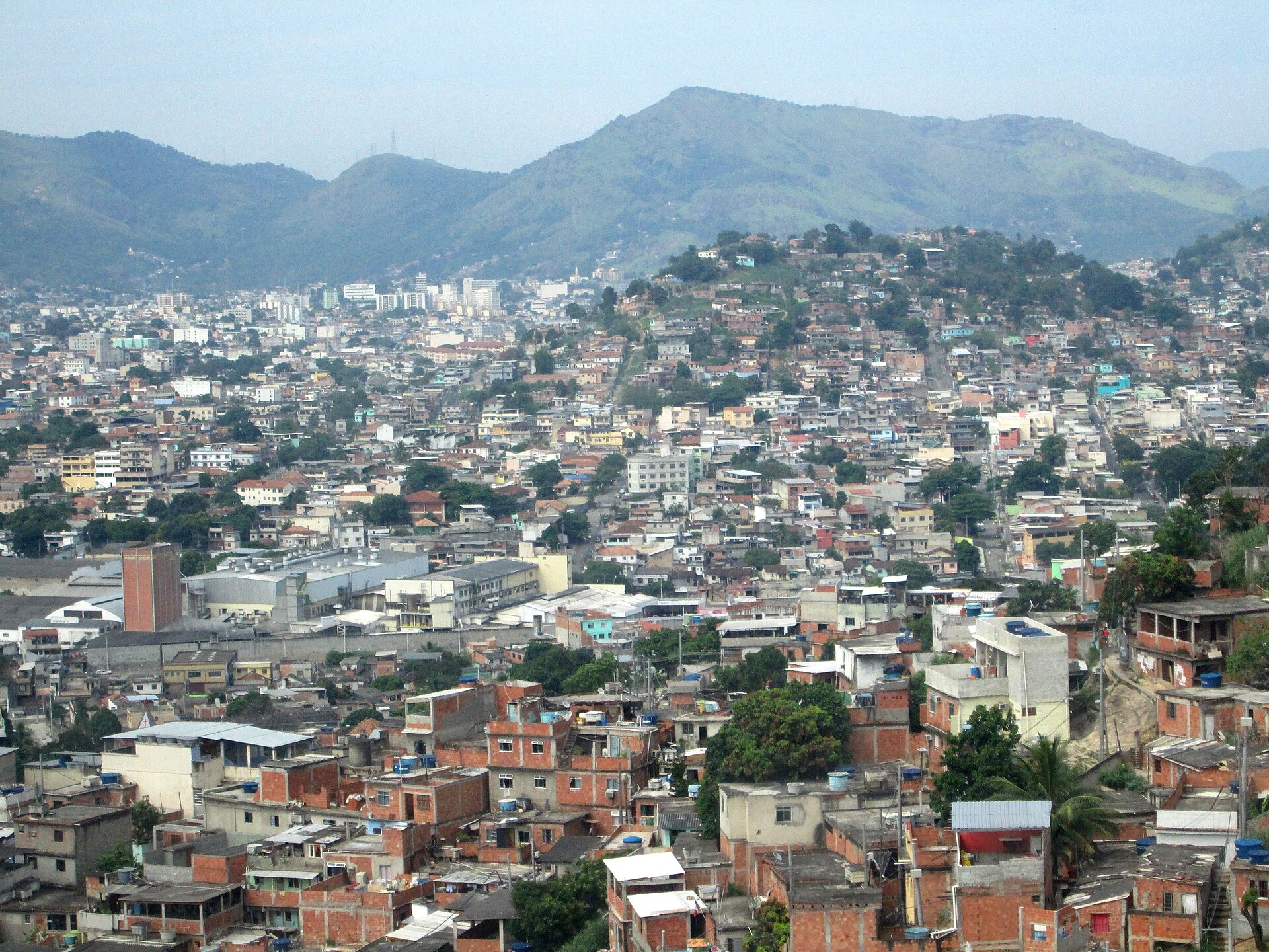 Hillside view of densely packed red-brick and concrete houses of the Complexo do Alemão favela in north Rio de Janeiro, with green hills and a hazy sky behind
