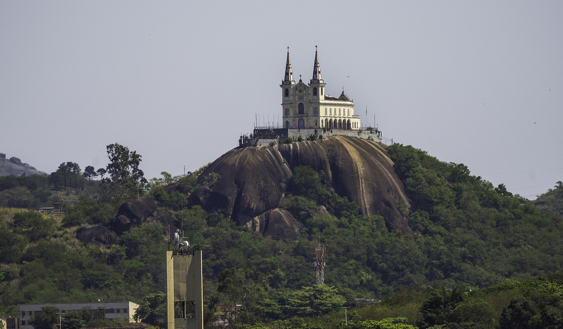 A white two-towered church on top of a large granite rock hill, with green trees below and a hazy grey sky