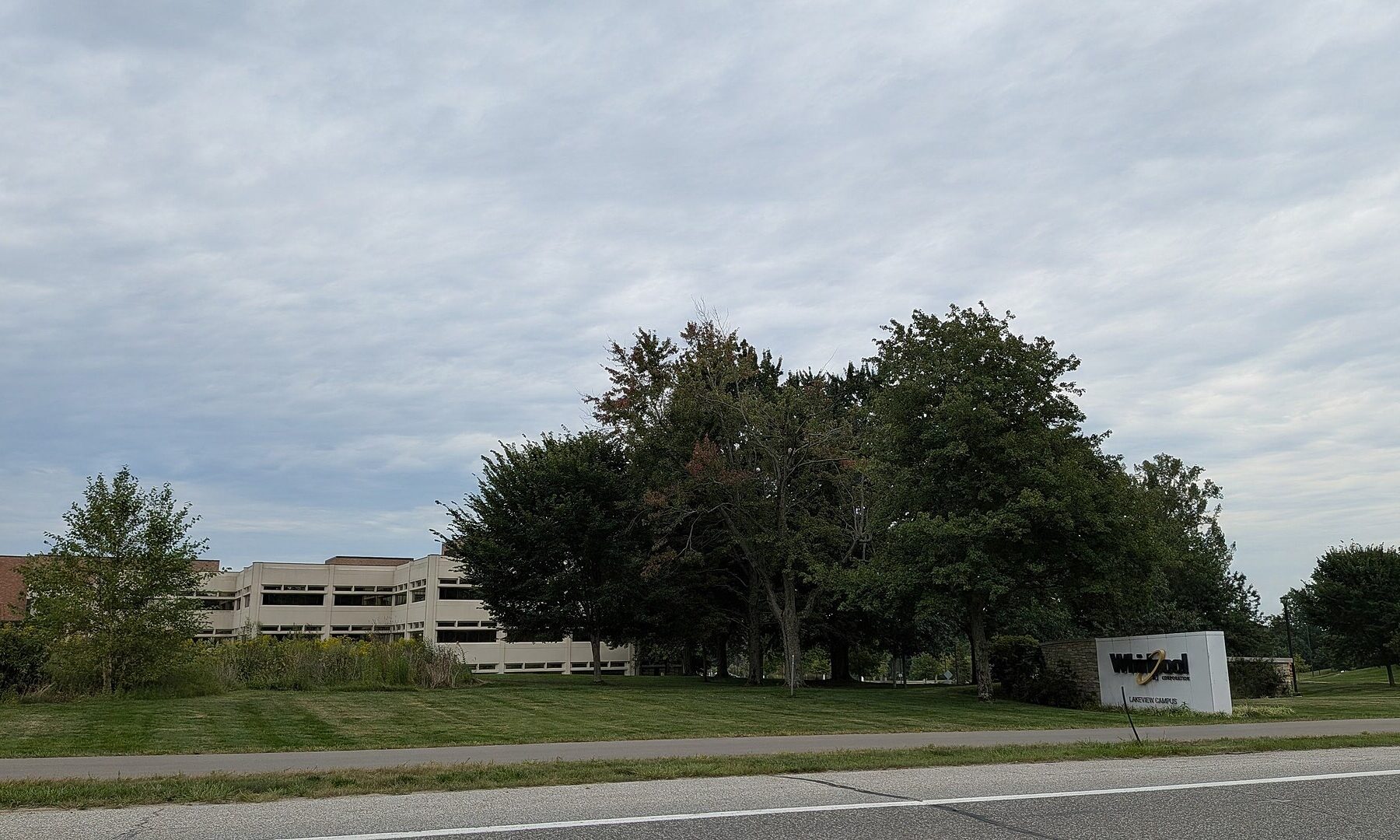 A low beige office building behind trees and a lawn, with a white Whirlpool sign at the roadside, under an overcast sky