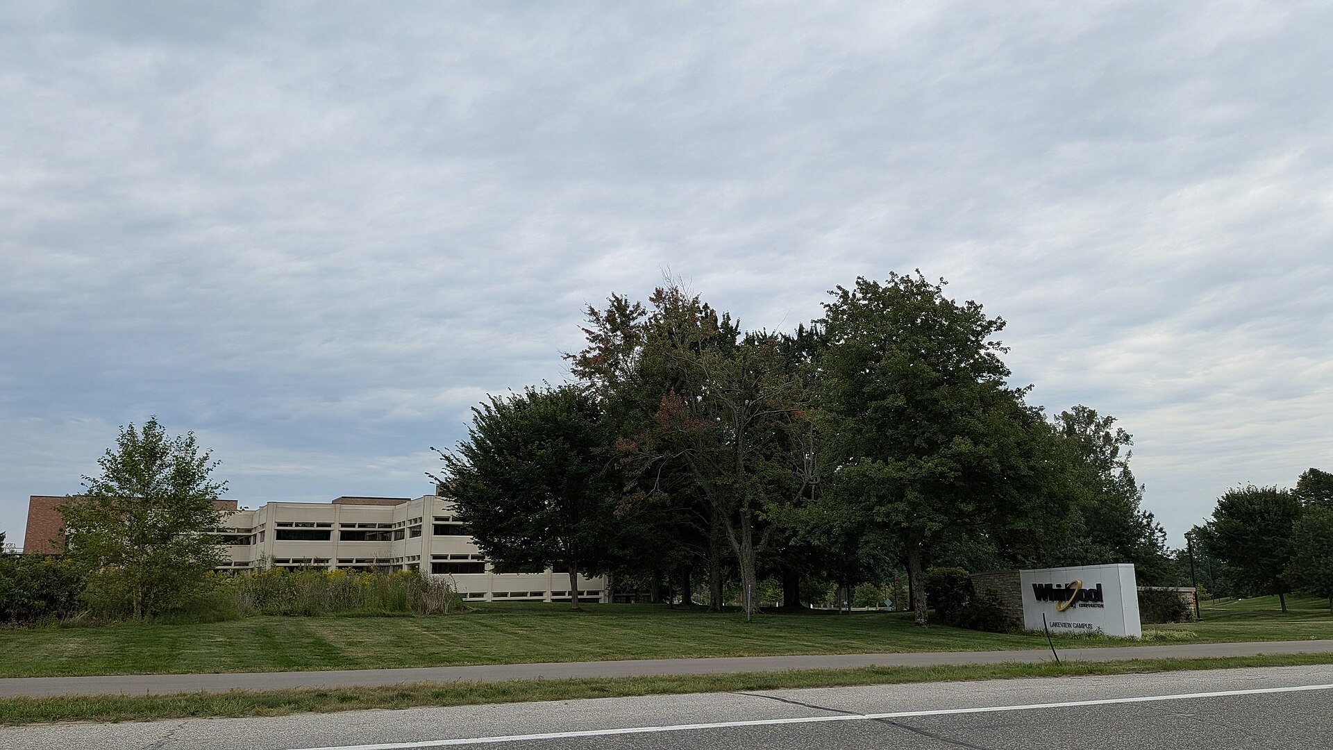 A low beige office building behind trees and a lawn, with a white Whirlpool sign at the roadside, under an overcast sky