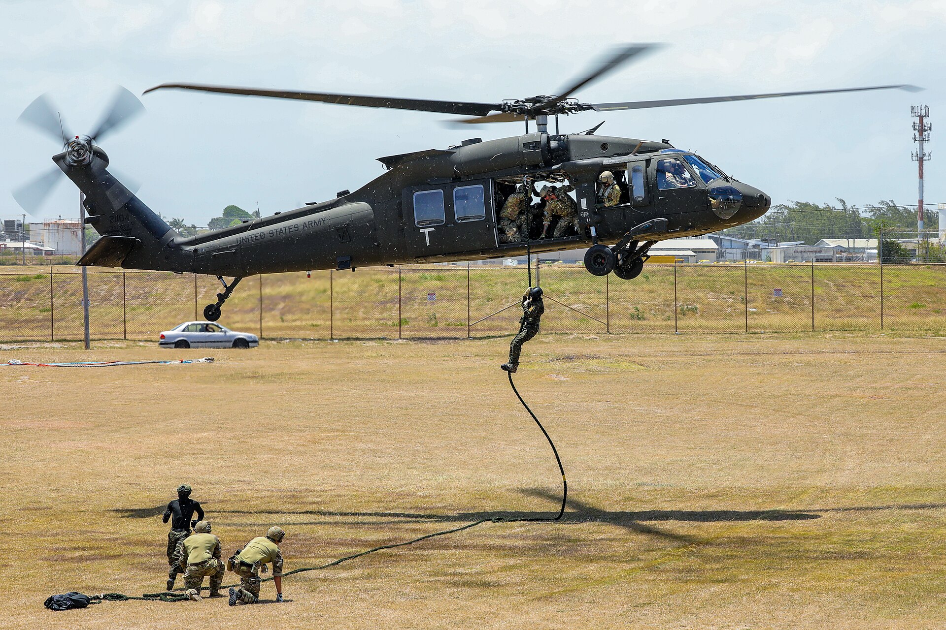 A US Army helicopter hovers while a soldier slides down a rope as three others kneel on dry grass below during a fast-rope drill