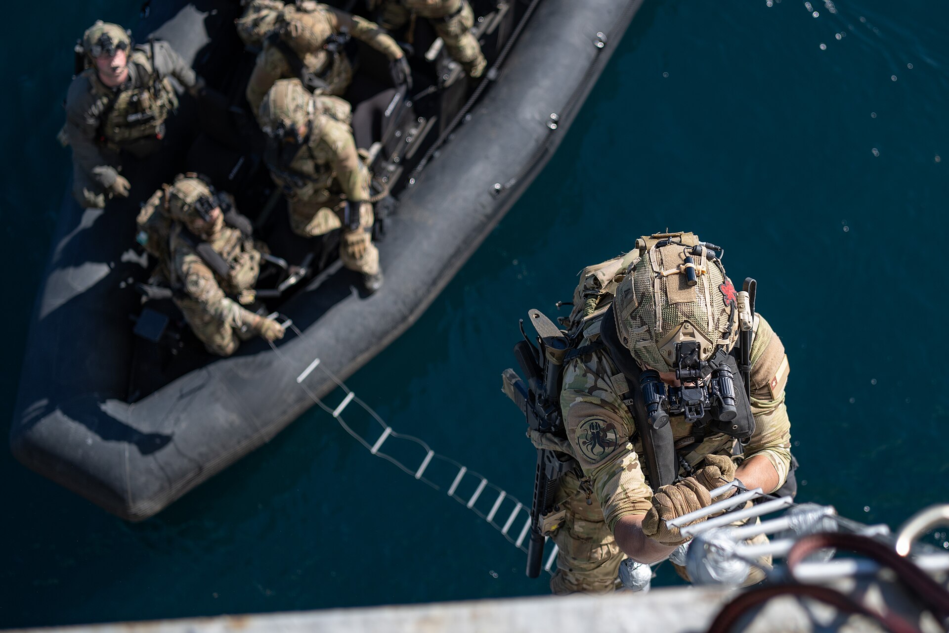 A Chilean Navy special forces operator in camouflage and helmet climbs a rope ladder up a ship's hull above an inflatable boat carrying other operators