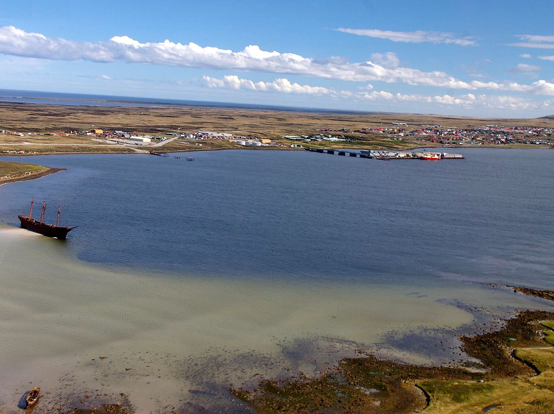 Aerial view of Stanley harbour in the Falkland Islands with a rusted shipwreck on the left, the town and waterfront across the bay and a cloudy blue sky
