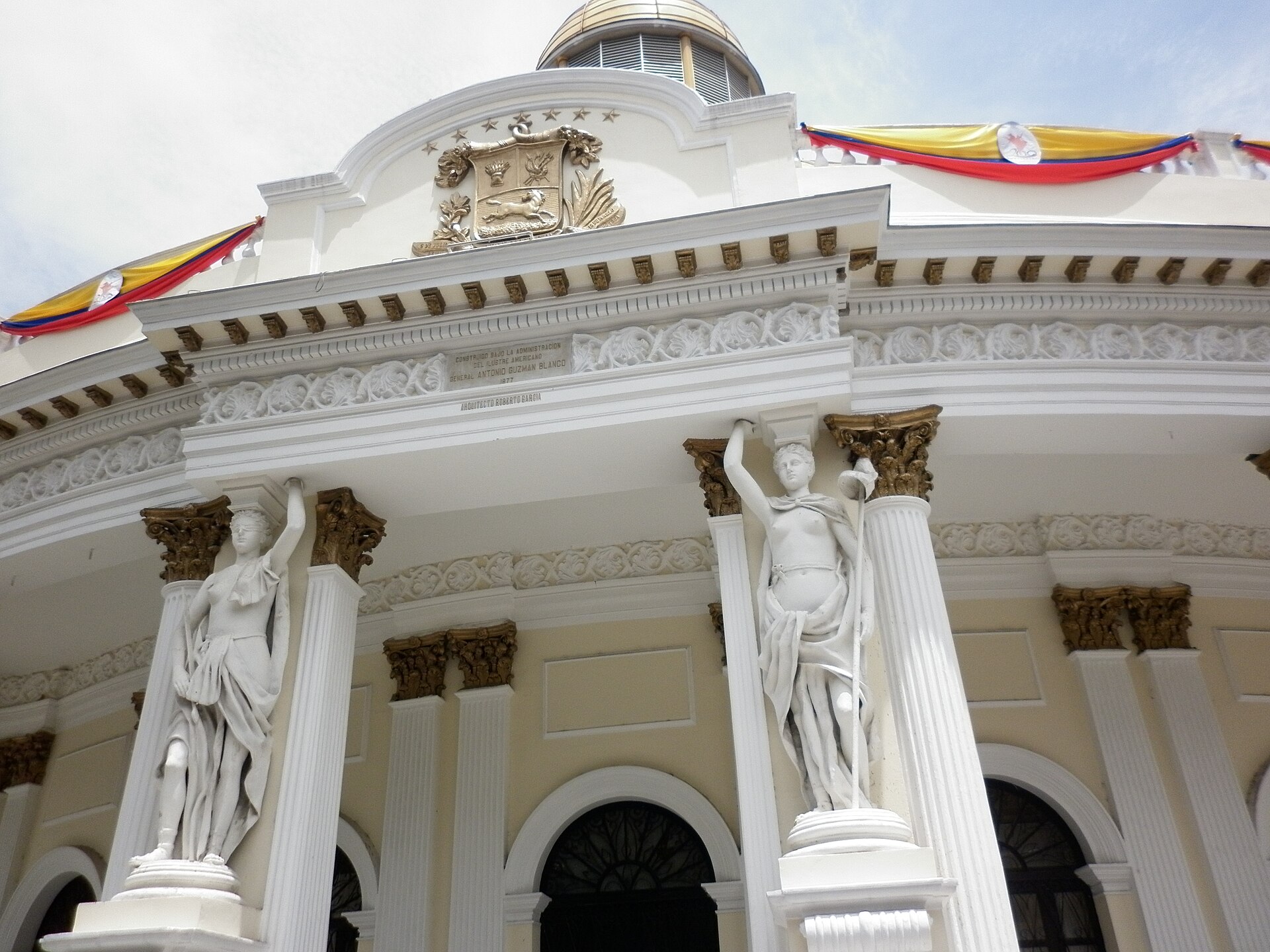 The curved cream facade of the Palacio Federal Legislativo in Caracas with white statues and columns, a golden dome and Venezuelan tricolour bunting