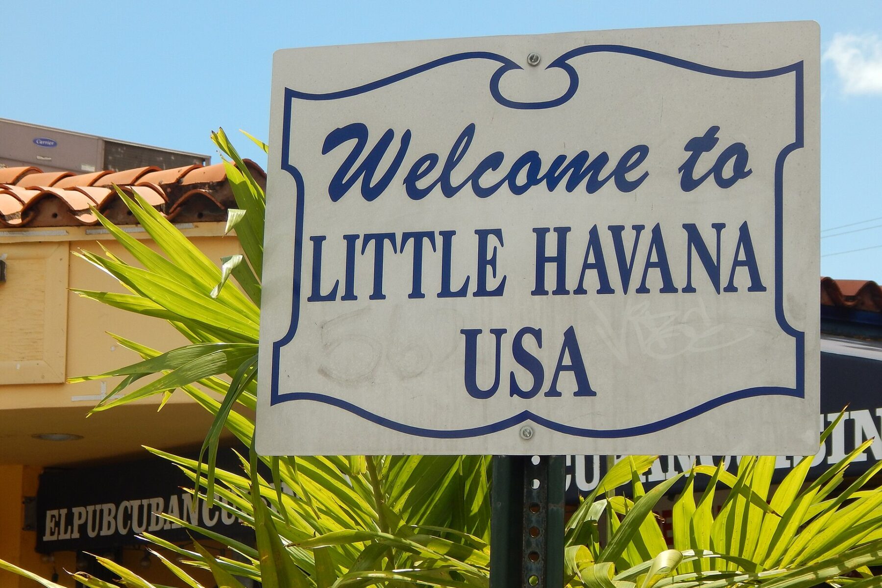 A white and blue 'Welcome to Little Havana USA' street sign among palm leaves, with a Cuban restaurant sign behind it