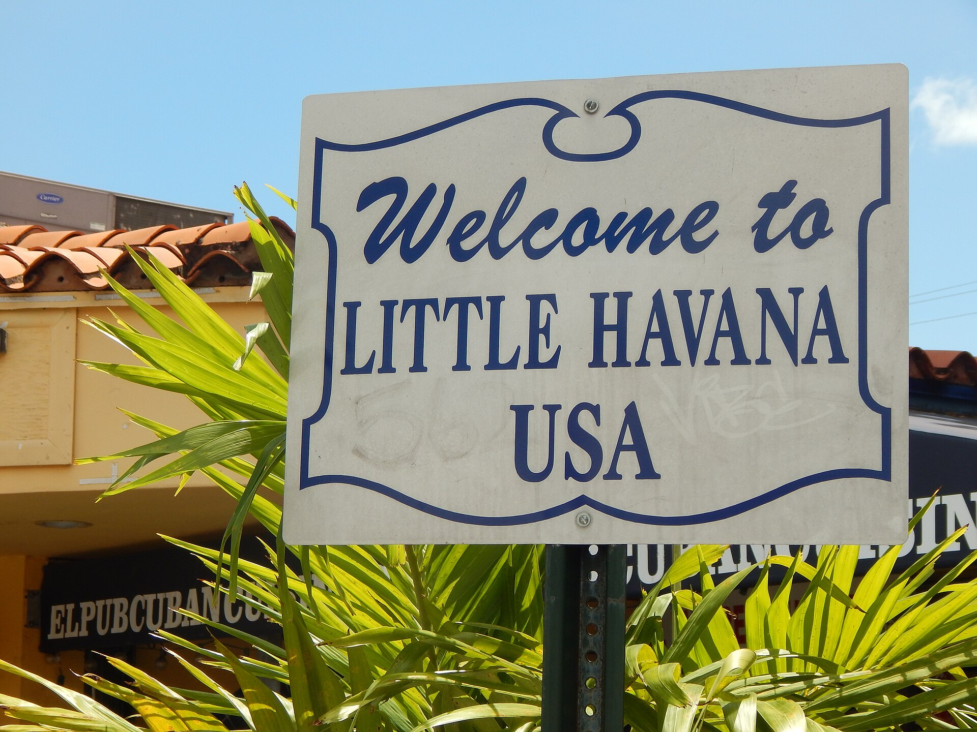 A white and blue 'Welcome to Little Havana USA' street sign among palm leaves, with a Cuban restaurant sign behind it