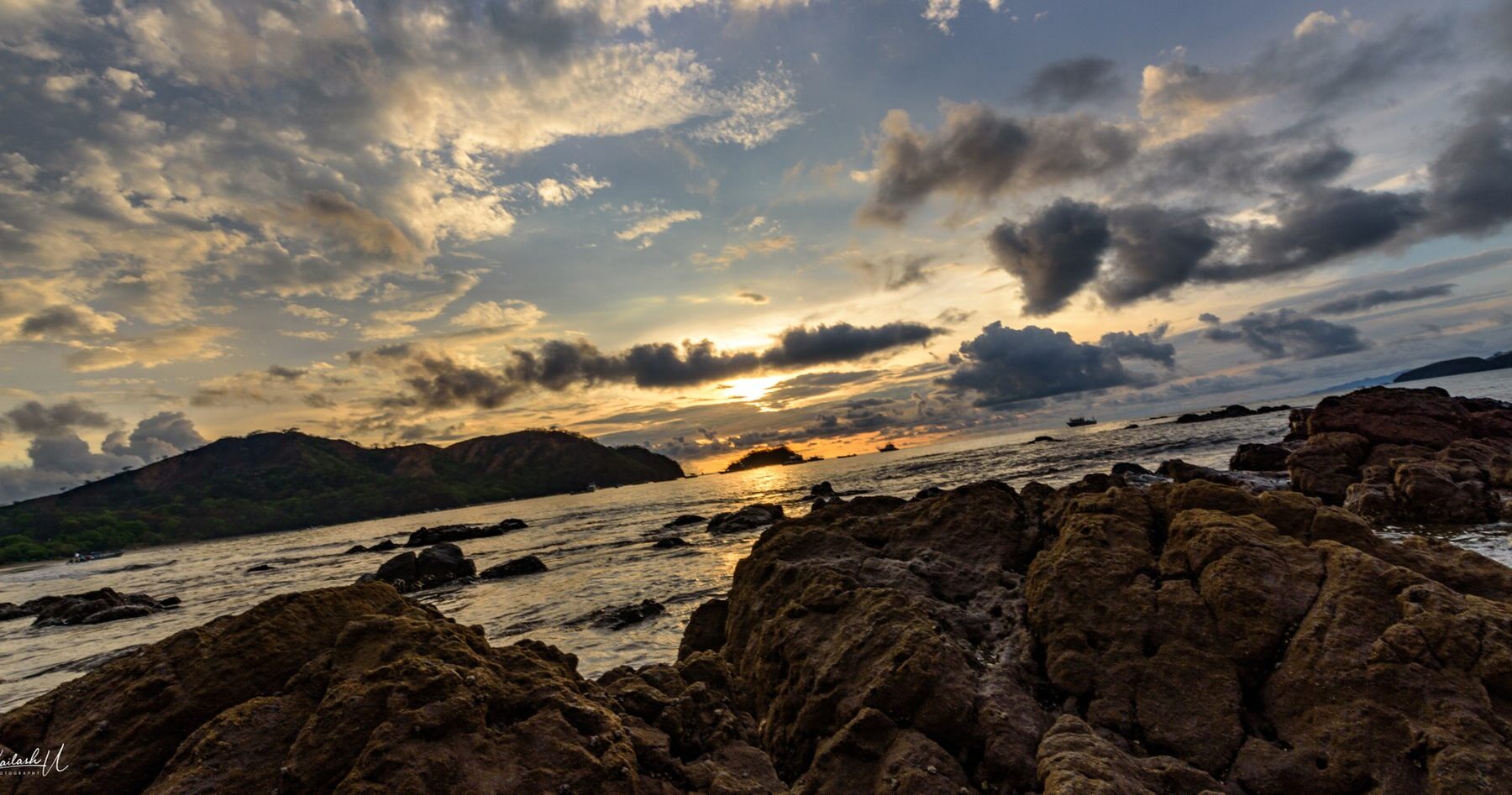 Dark volcanic rocks in the foreground of a bay at Playas del Coco, with green headlands and a cloudy sky lit by the setting sun