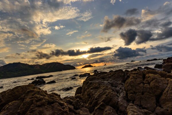 Dark volcanic rocks in the foreground of a bay at Playas del Coco, with green headlands and a cloudy sky lit by the setting sun