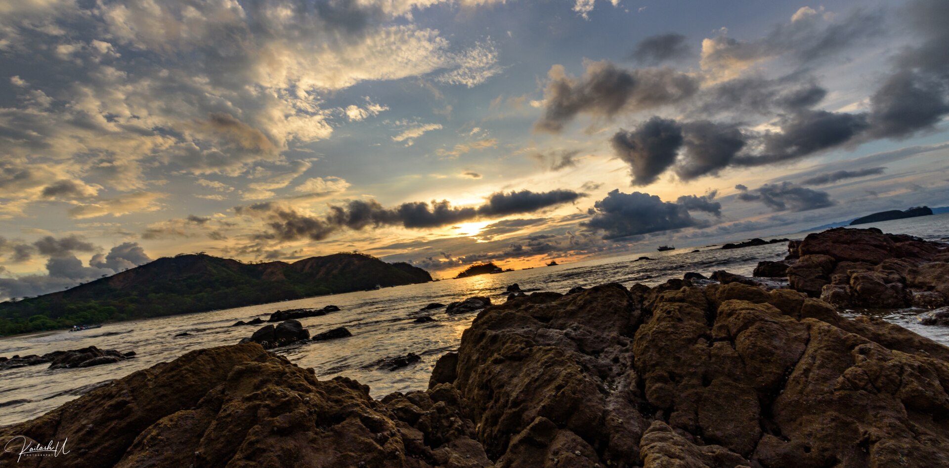 Dark volcanic rocks in the foreground of a bay at Playas del Coco, with green headlands and a cloudy sky lit by the setting sun
