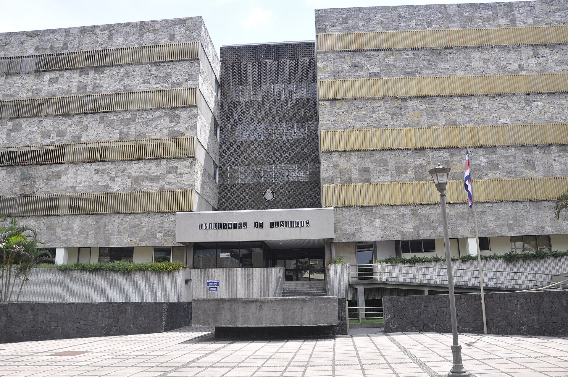 The stone-clad Tribunales de Justicia building in San José with its entrance sign, a Costa Rican flag and a paved plaza in front
