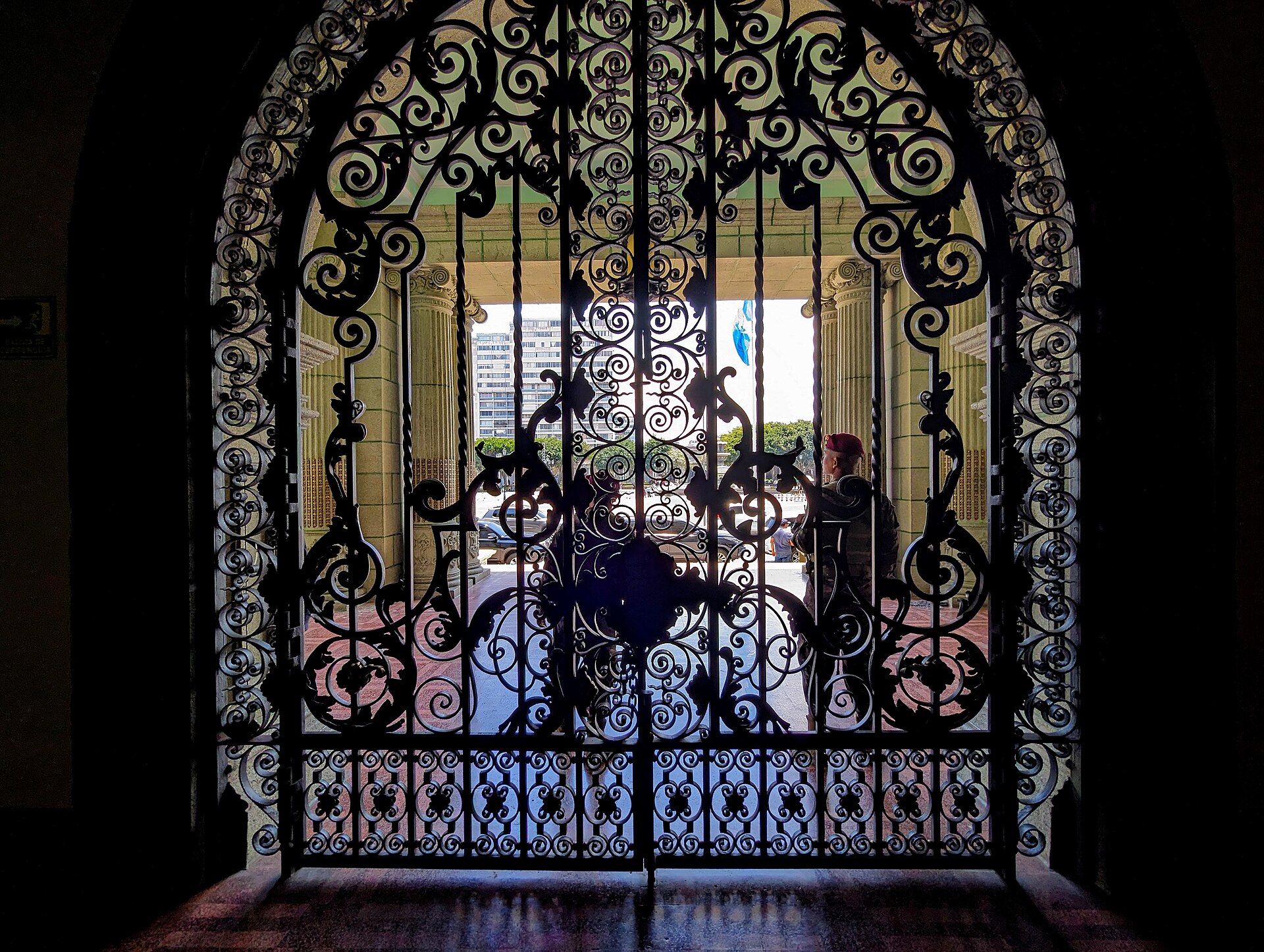 A black wrought-iron gate with scrollwork at the National Palace of Culture in Guatemala City, a soldier standing beyond it