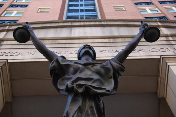 Bronze statue of Lady Justice with arms raised holding scales beneath the entrance of a federal courthouse with a brick facade