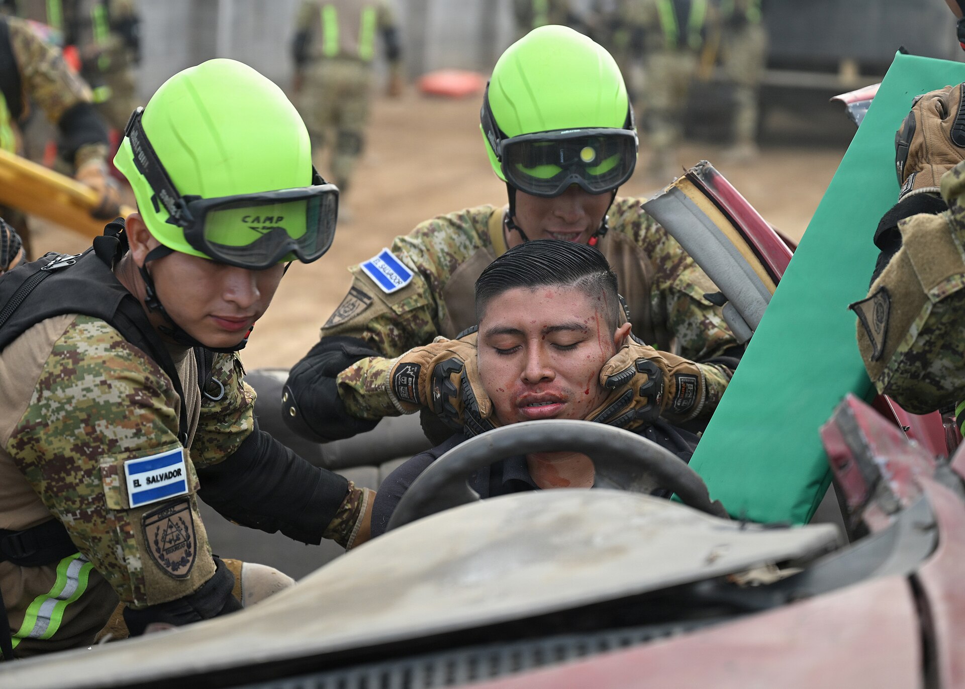 Three Salvadoran soldiers in camouflage, two in lime-green helmets and goggles, give first aid to a volunteer playing a trapped victim in a wrecked car