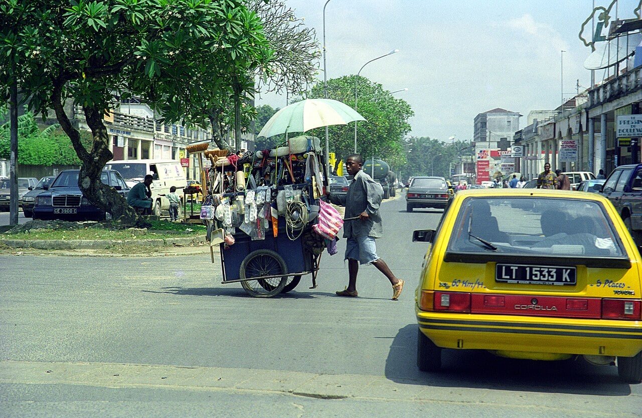 A street vendor pushes a loaded cart across a busy road in Douala, Cameroon, with a yellow taxi in the foreground