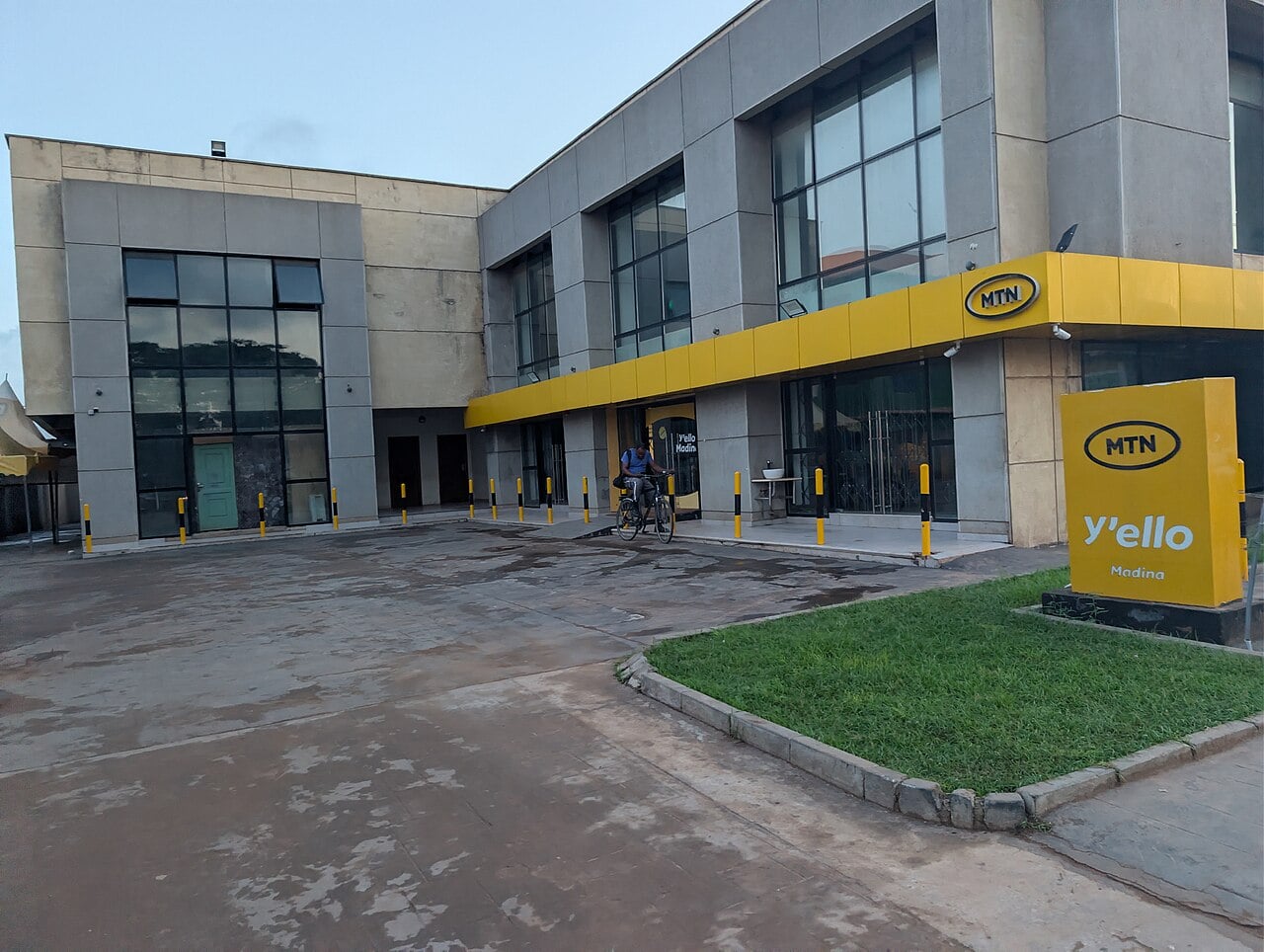 A grey two-storey building with a yellow MTN band and a yellow MTN Y'ello Madina sign, with a man on a bicycle at the entrance