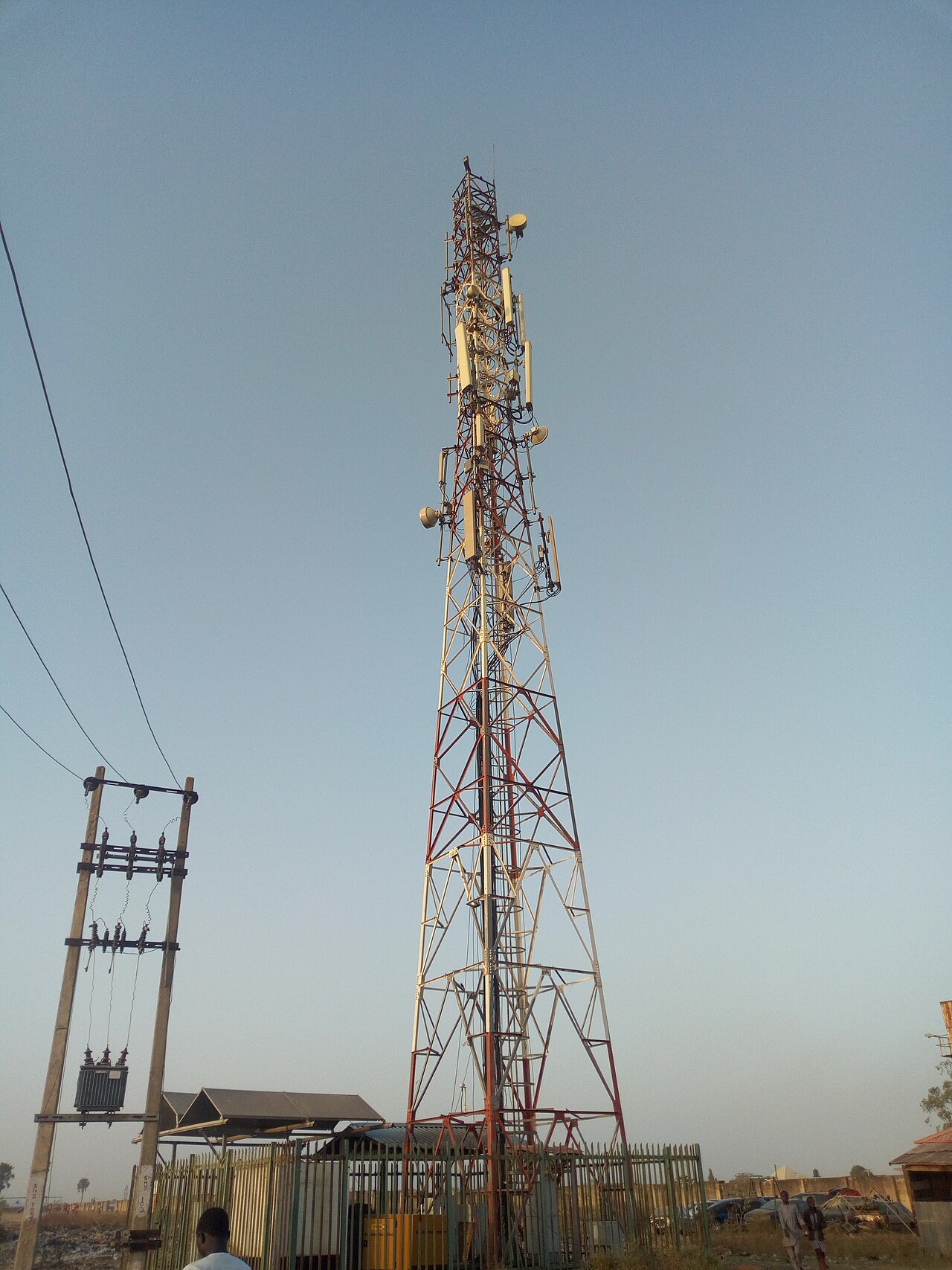 A tall steel telecom mast with antennas and microwave dishes beside a power transformer in Kaduna State, Nigeria