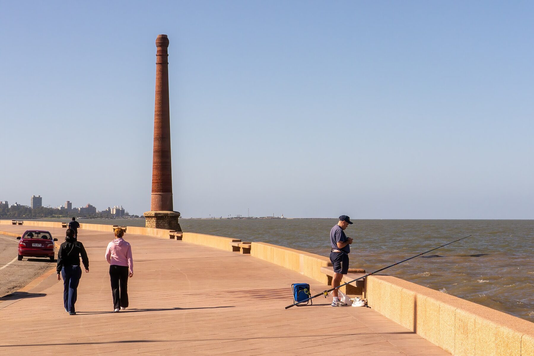 People walk and fish along the Rambla seafront in Montevideo beside the Río de la Plata, with an old brick chimney