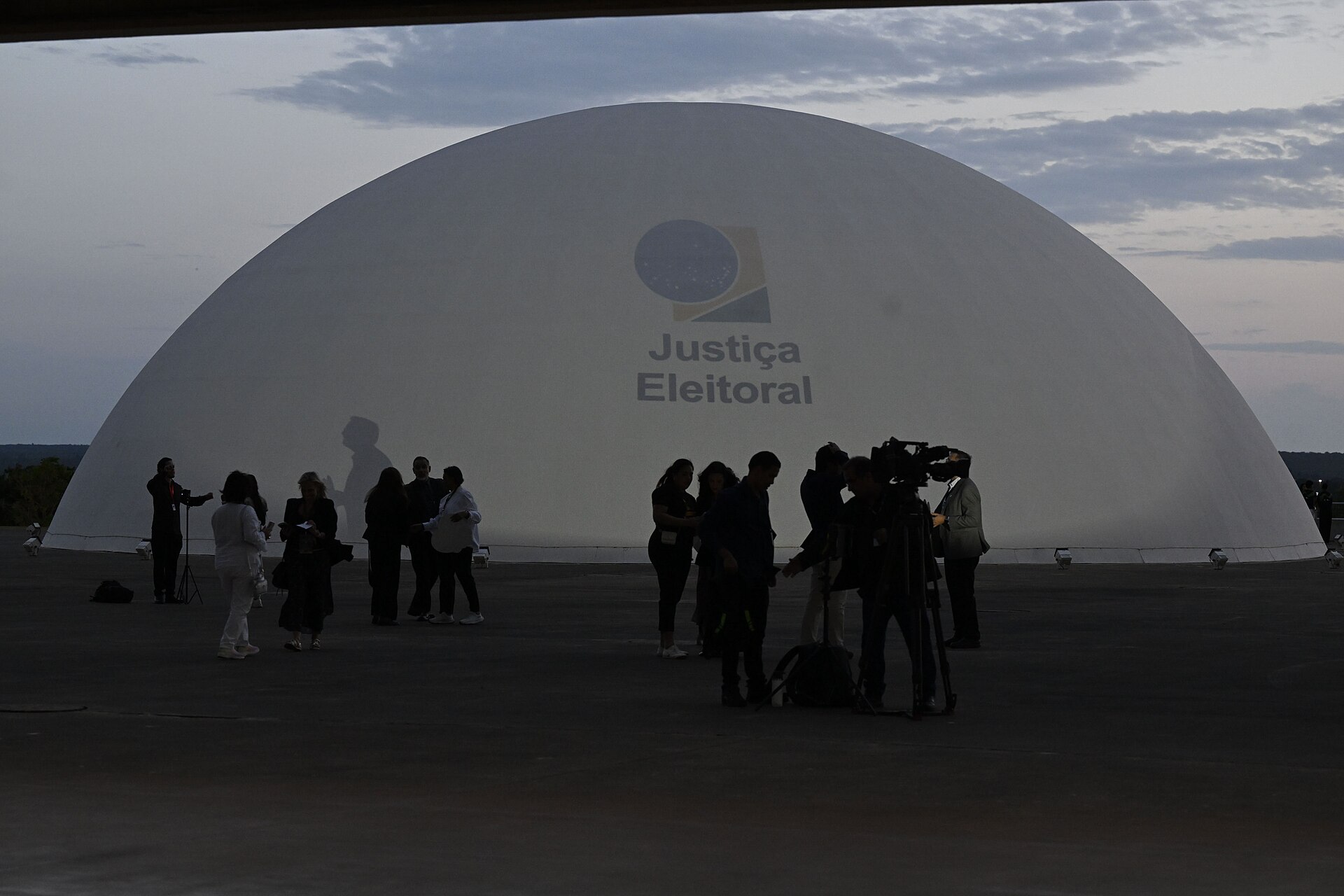 People and a TV crew in silhouette in front of a large white dome lit with the Justiça Eleitoral logo at dusk