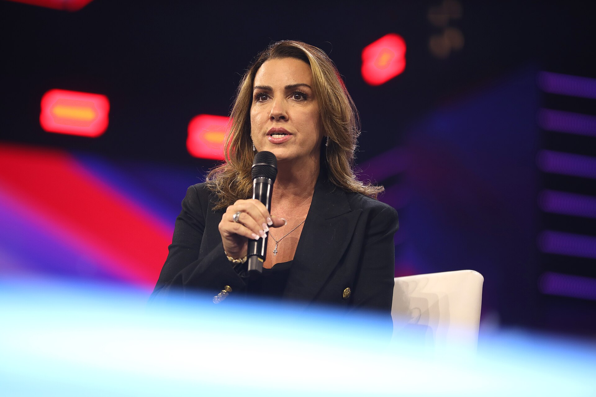 Sara Carter, seated in a dark blazer, speaks into a handheld microphone on a stage lit in red and purple