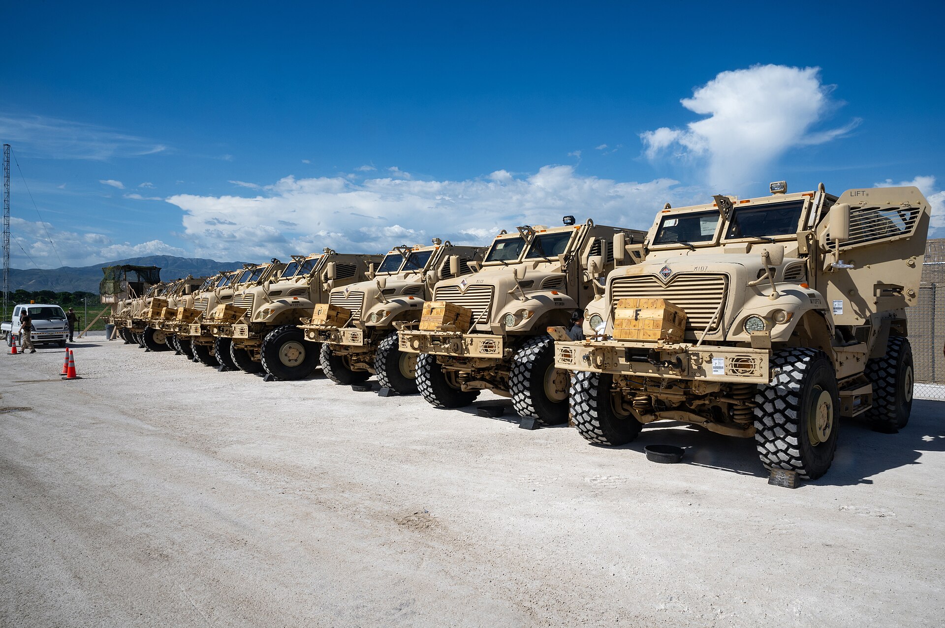 A row of sand-coloured armoured MRAP vehicles parked on a gravel apron under a blue sky, with hills behind