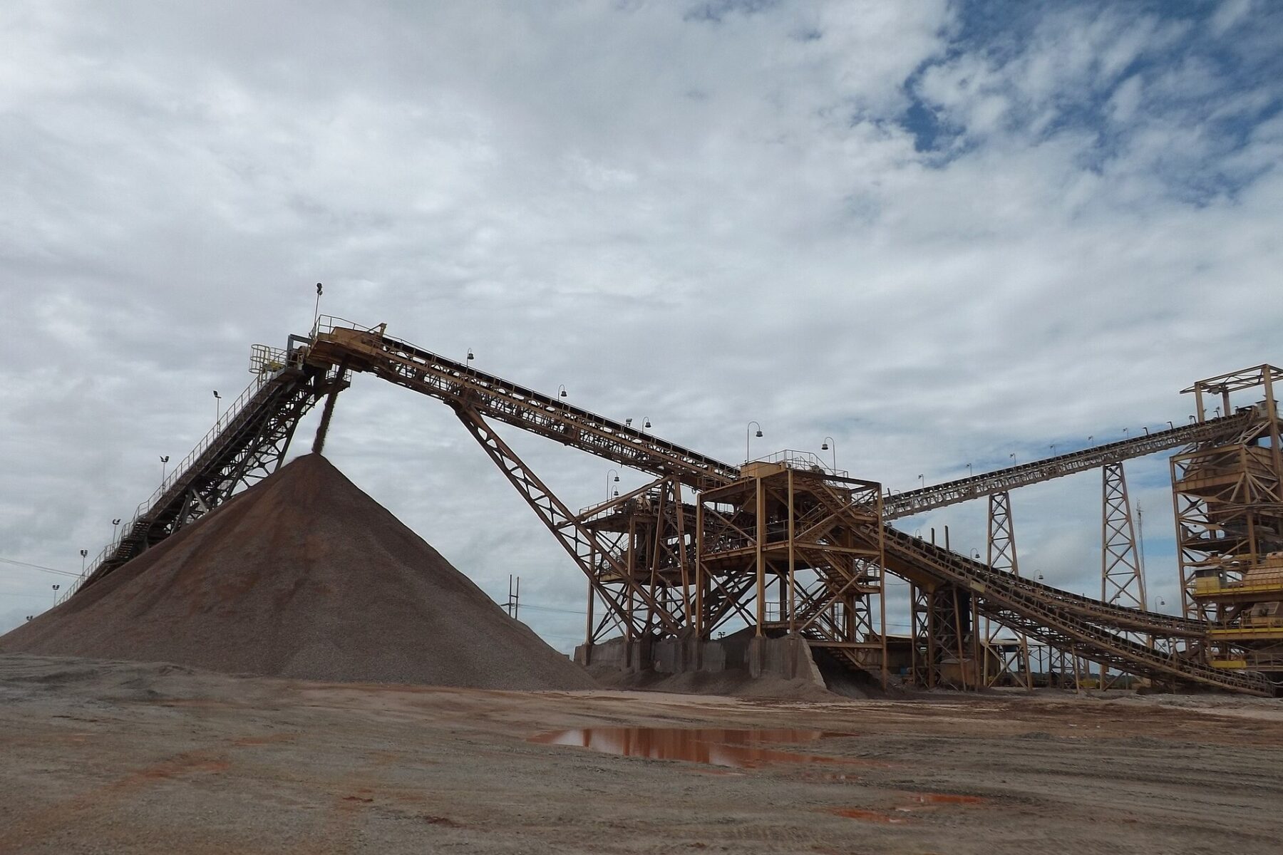 Conveyor belts and steel towers feeding a large pile of crushed ore at a mine processing plant under a cloudy sky