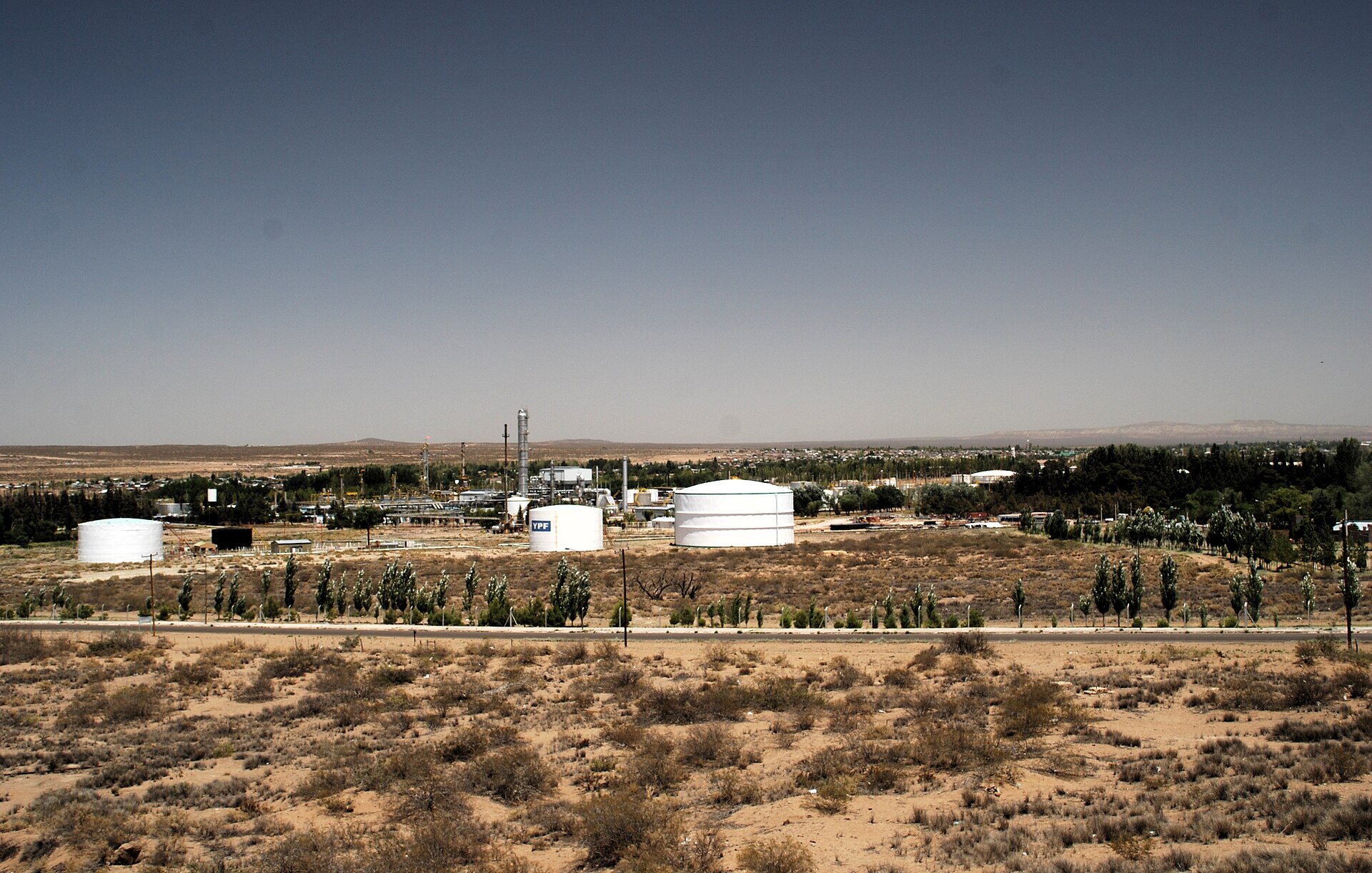 White oil storage tanks and refinery towers on a dry, scrubby plain under a clear sky