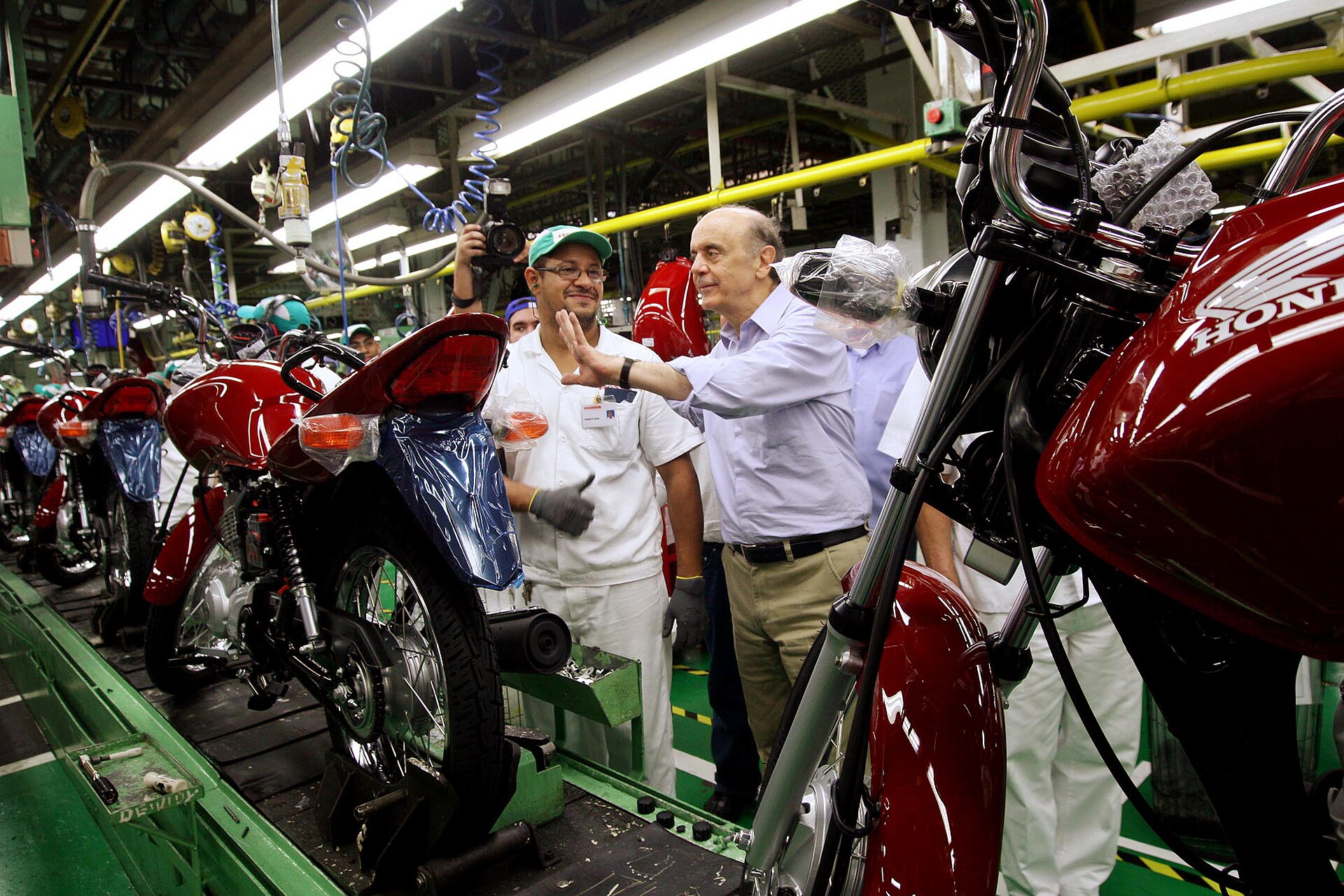Red Honda motorcycles on an assembly line with workers in white uniforms and a grey-haired man in a blue shirt gesturing