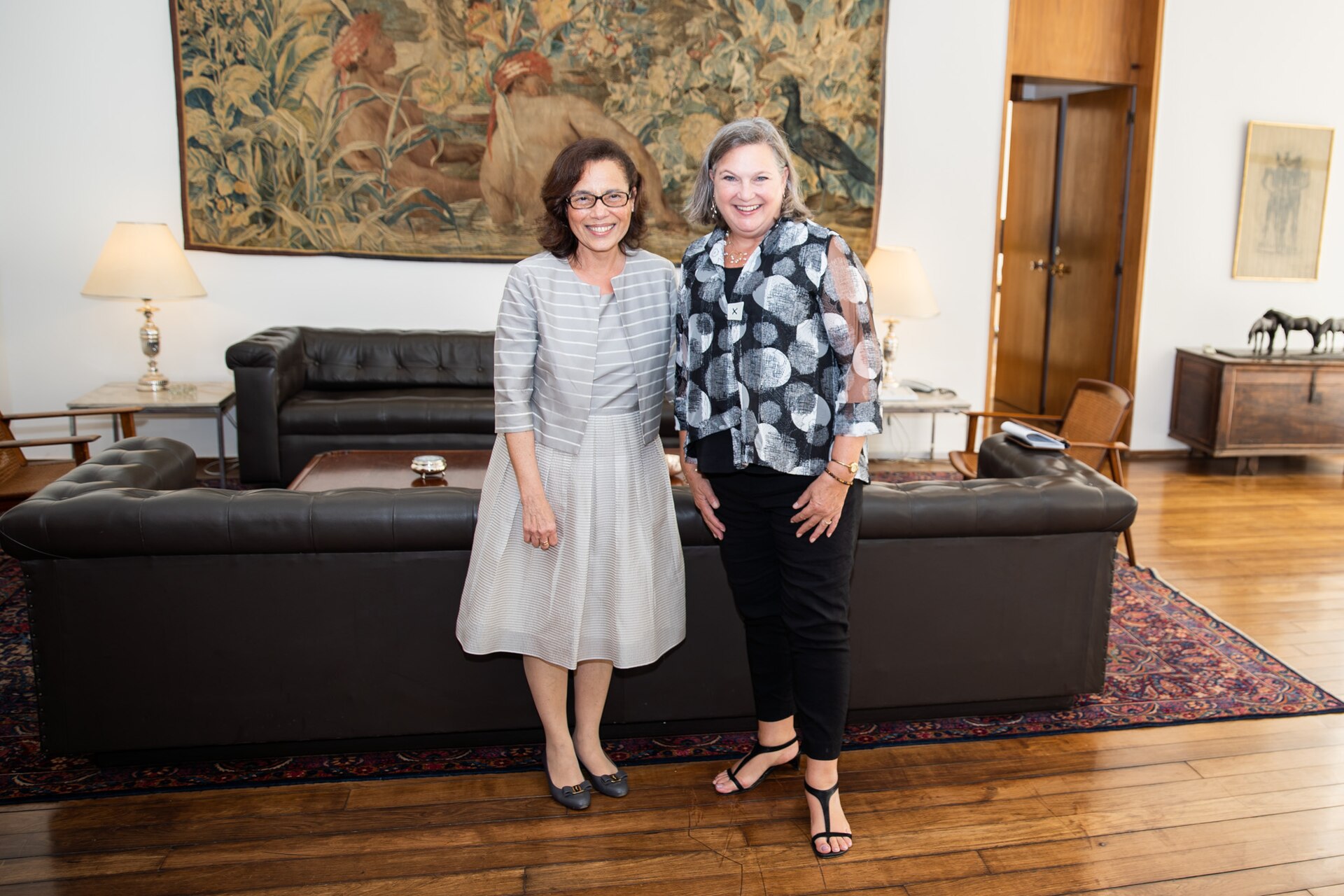 Two women standing in front of a black leather sofa beneath a large tapestry in a wood-floored reception room