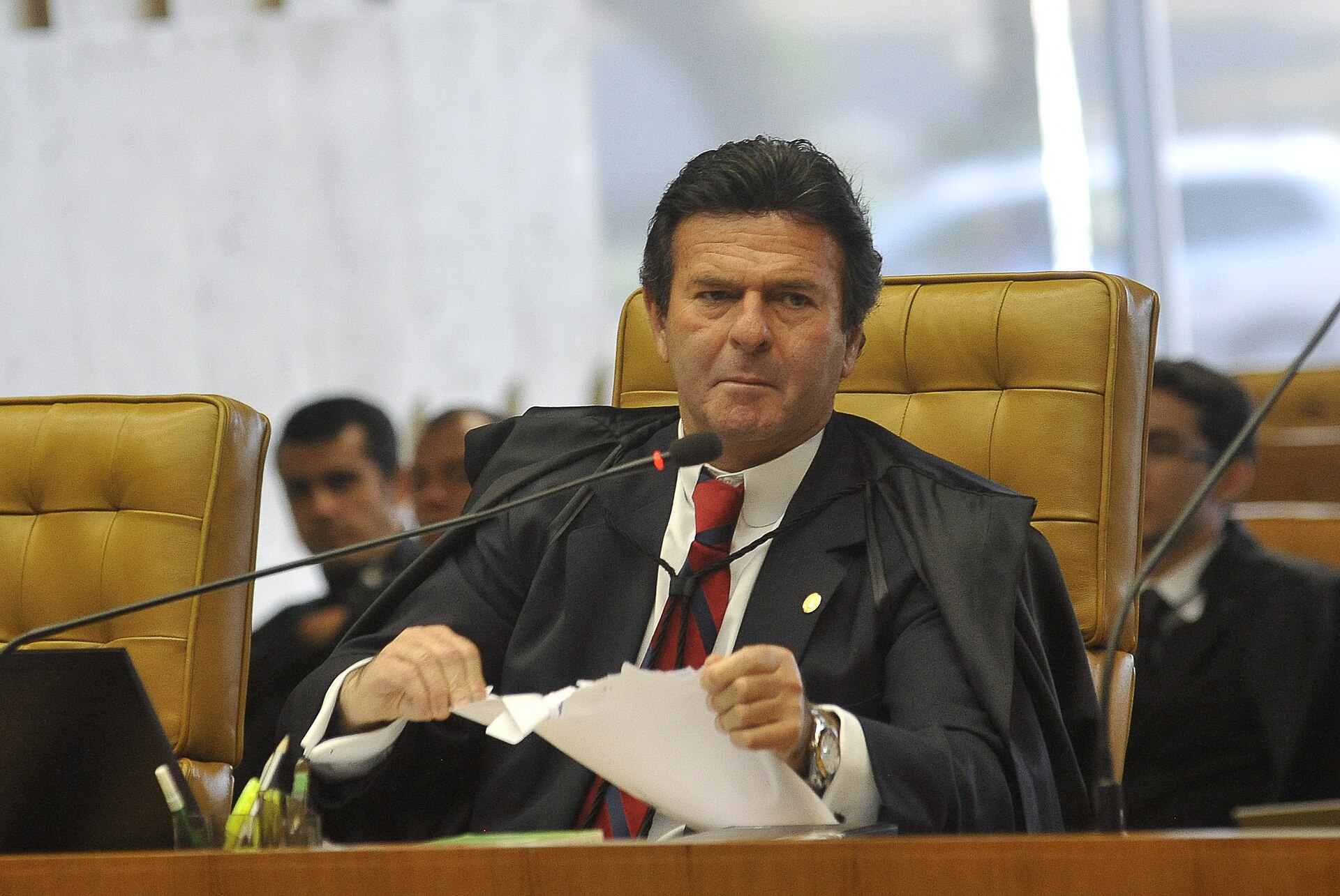 Justice Luiz Fux in a black robe and red striped tie, holding papers at the bench in the Supreme Court plenary
