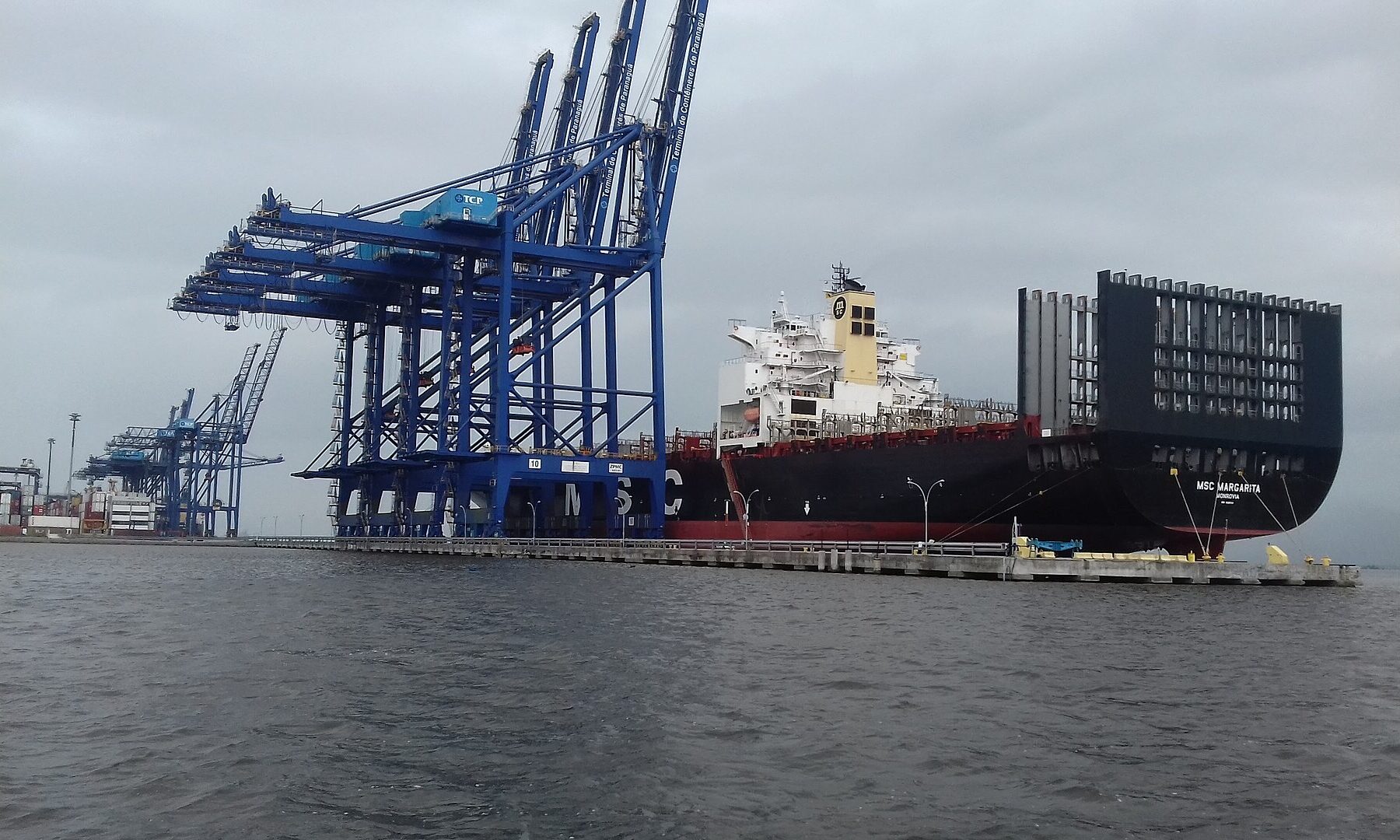 A large container ship moored beside blue ship-to-shore cranes at a container terminal under a grey sky