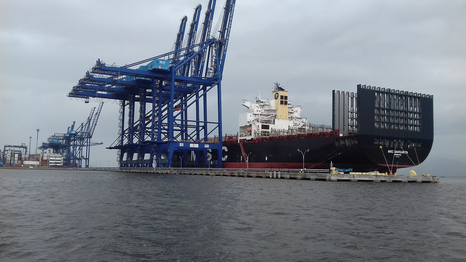 A large container ship moored beside blue ship-to-shore cranes at a container terminal under a grey sky