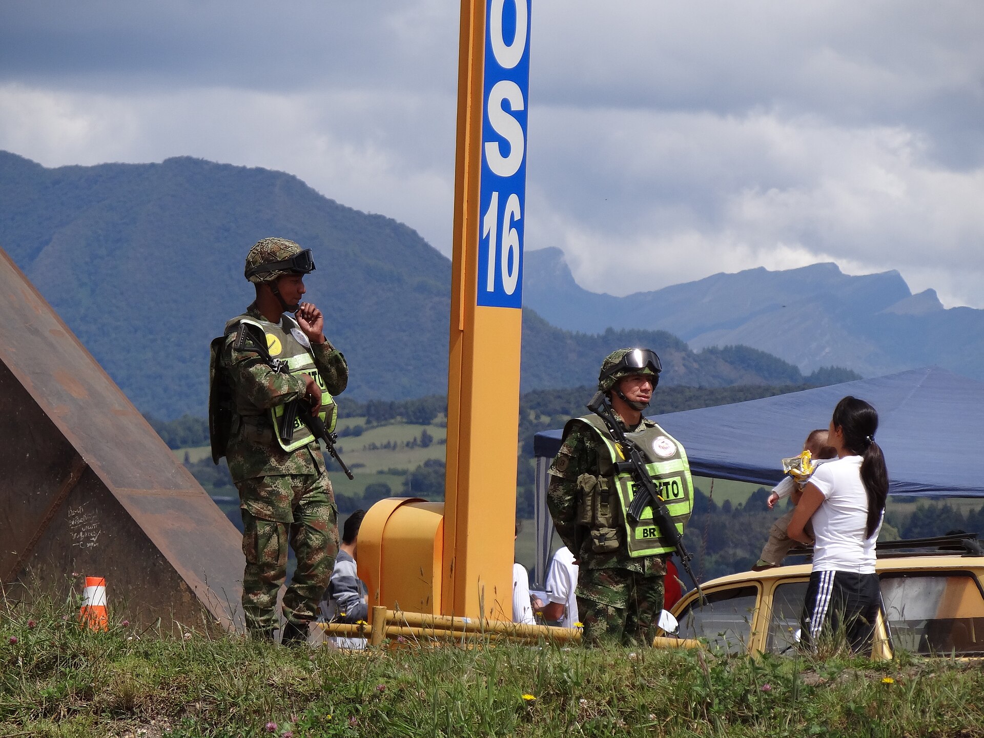 Two Colombian Army soldiers in camouflage and green vests with rifles stand beside a road, with mountains behind