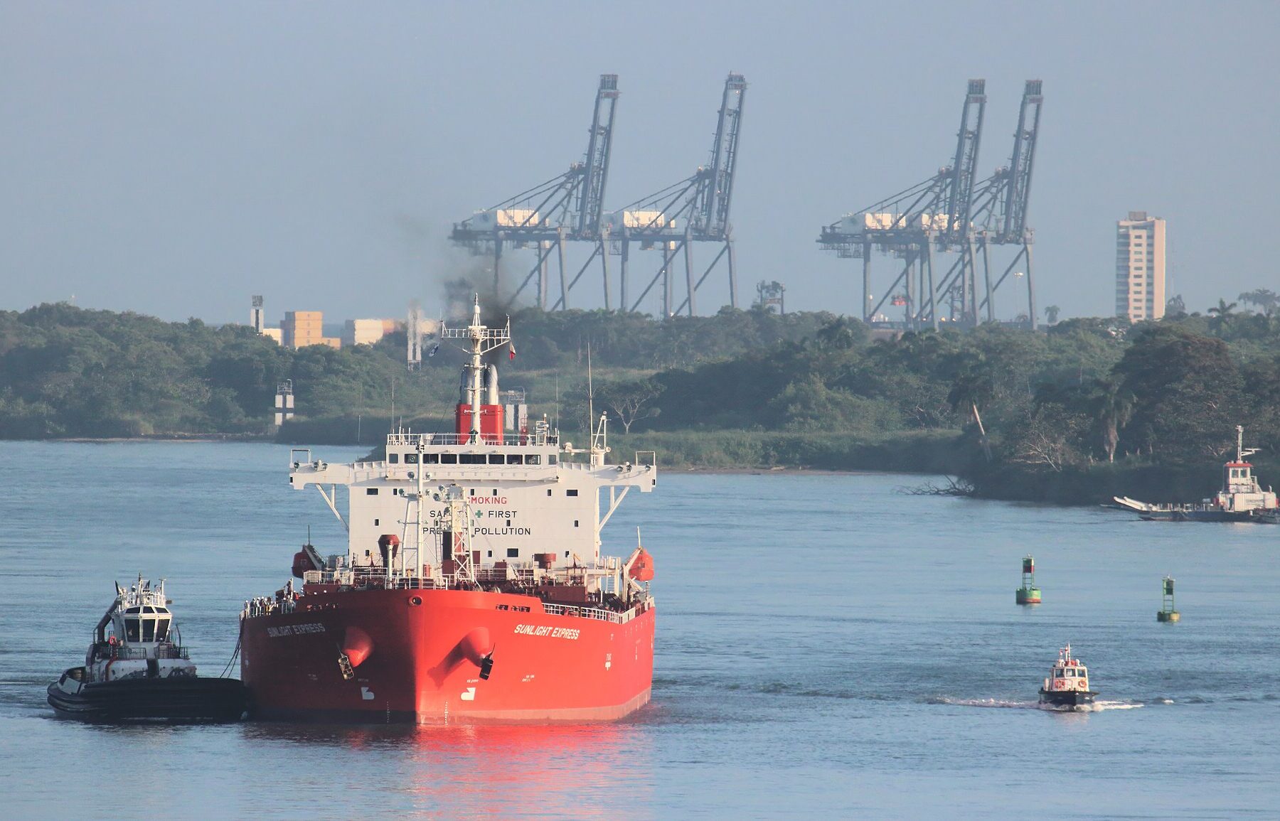 A red oil tanker escorted by tugboats in the Panama Canal's Caribbean entrance near Colón, with container cranes behind