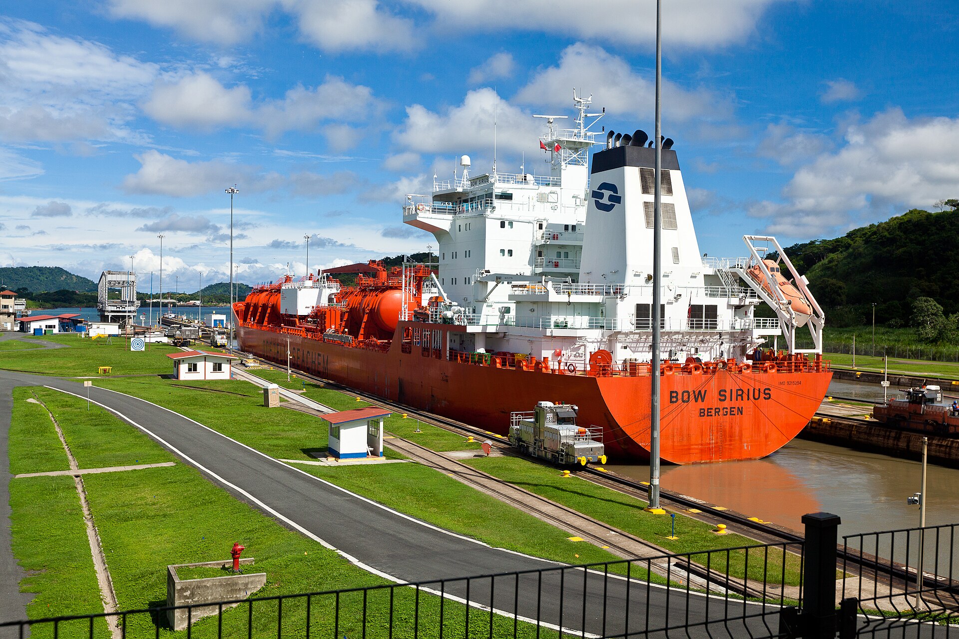 An orange oil and chemical tanker inside a Panama Canal lock beside a green lawn, a road and a locomotive track