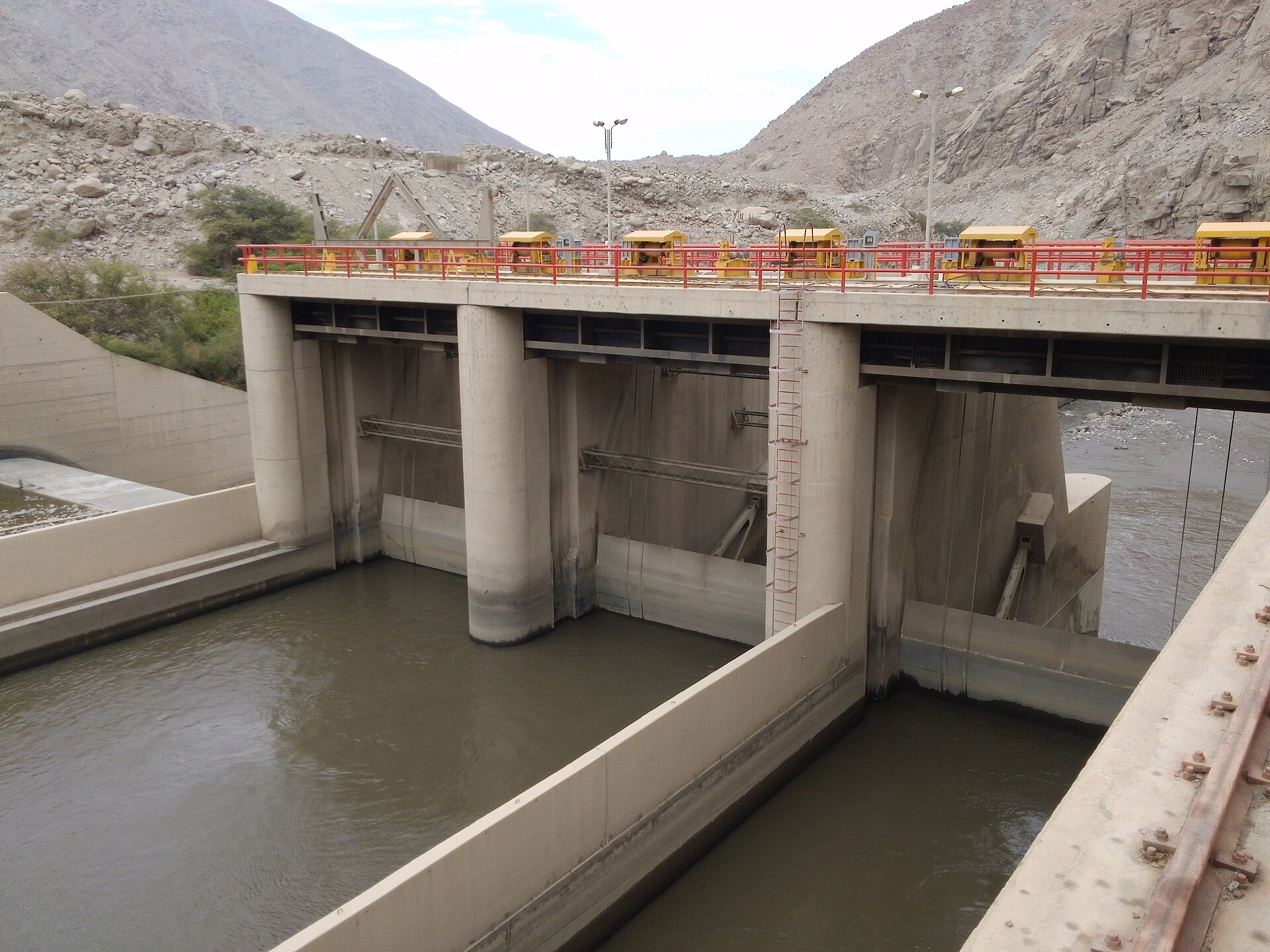 A concrete water intake structure with gates and a red-railed walkway spanning a brown river channel between dry rocky hills