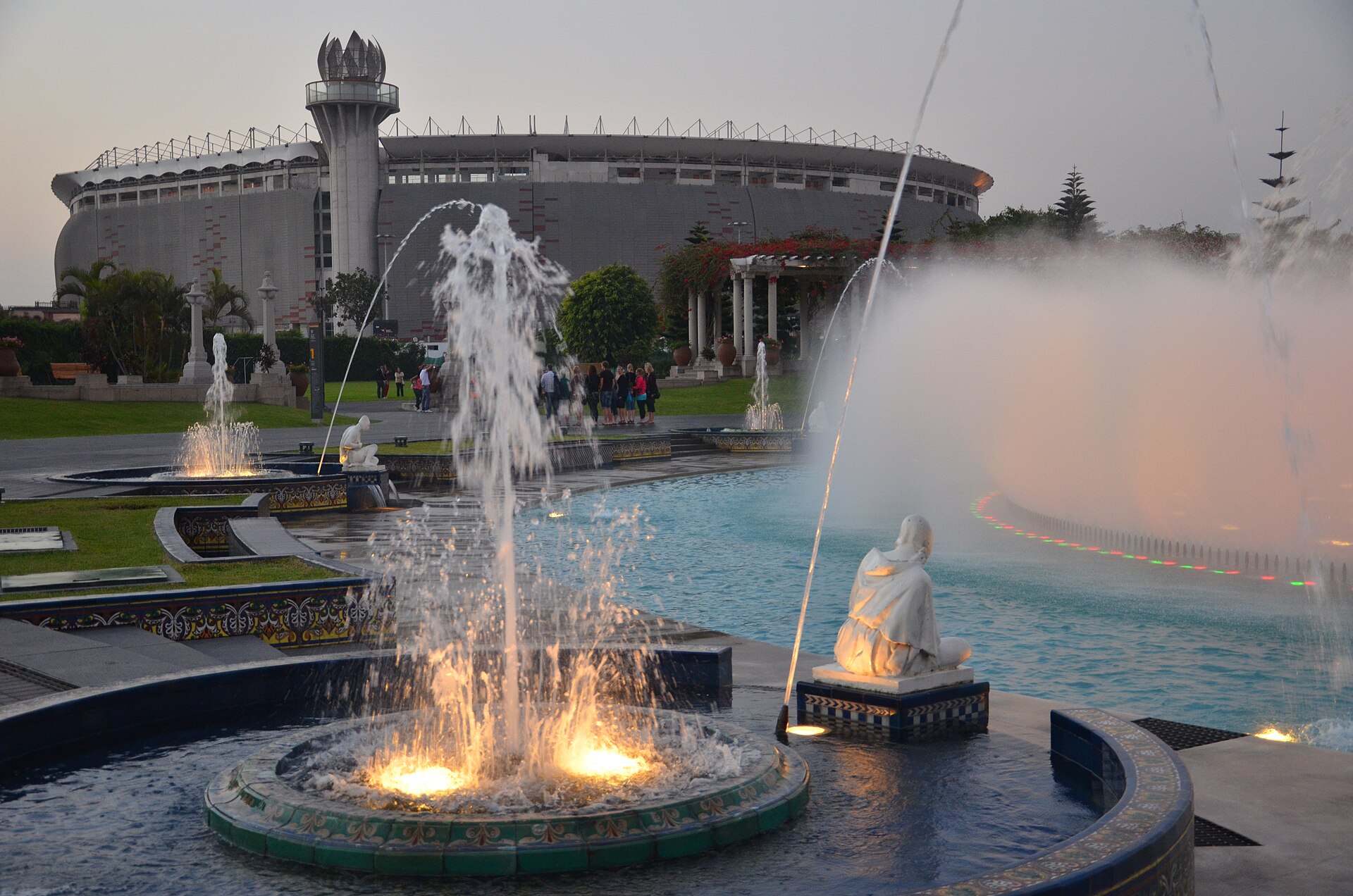 Illuminated fountains and white statues in Lima's Parque de la Reserva at dusk, with the Estadio Nacional and its tower behind