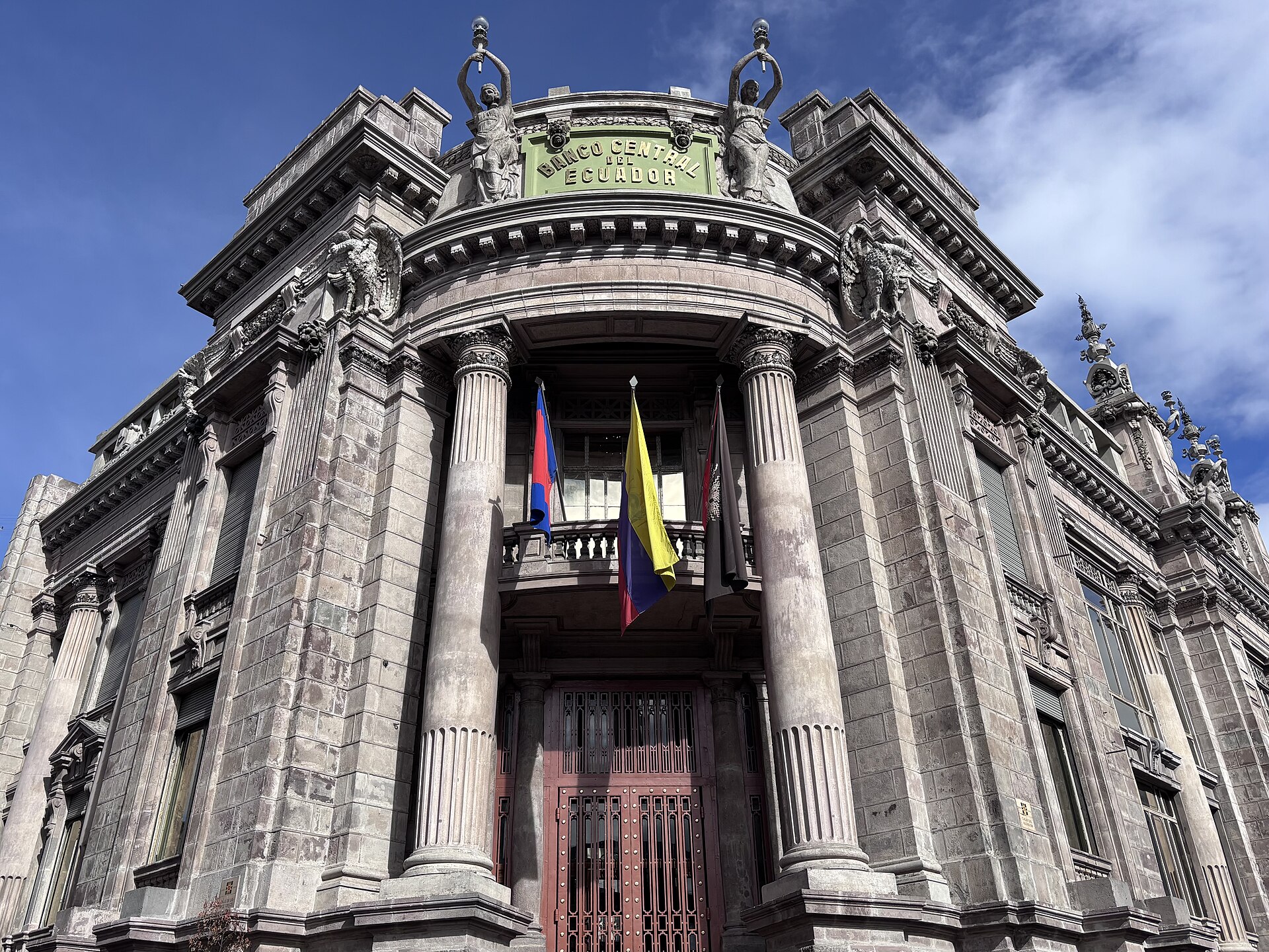 Ornate grey stone facade with columns, statues and a green sign reading Banco Central del Ecuador, with flags on a balcony under blue sky